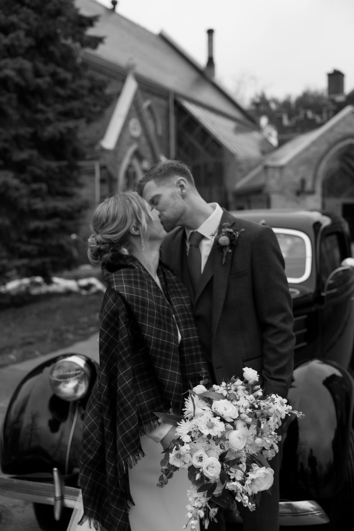 A romantic black and white wedding photo series featuring a couple with white flowers against a vintage car.