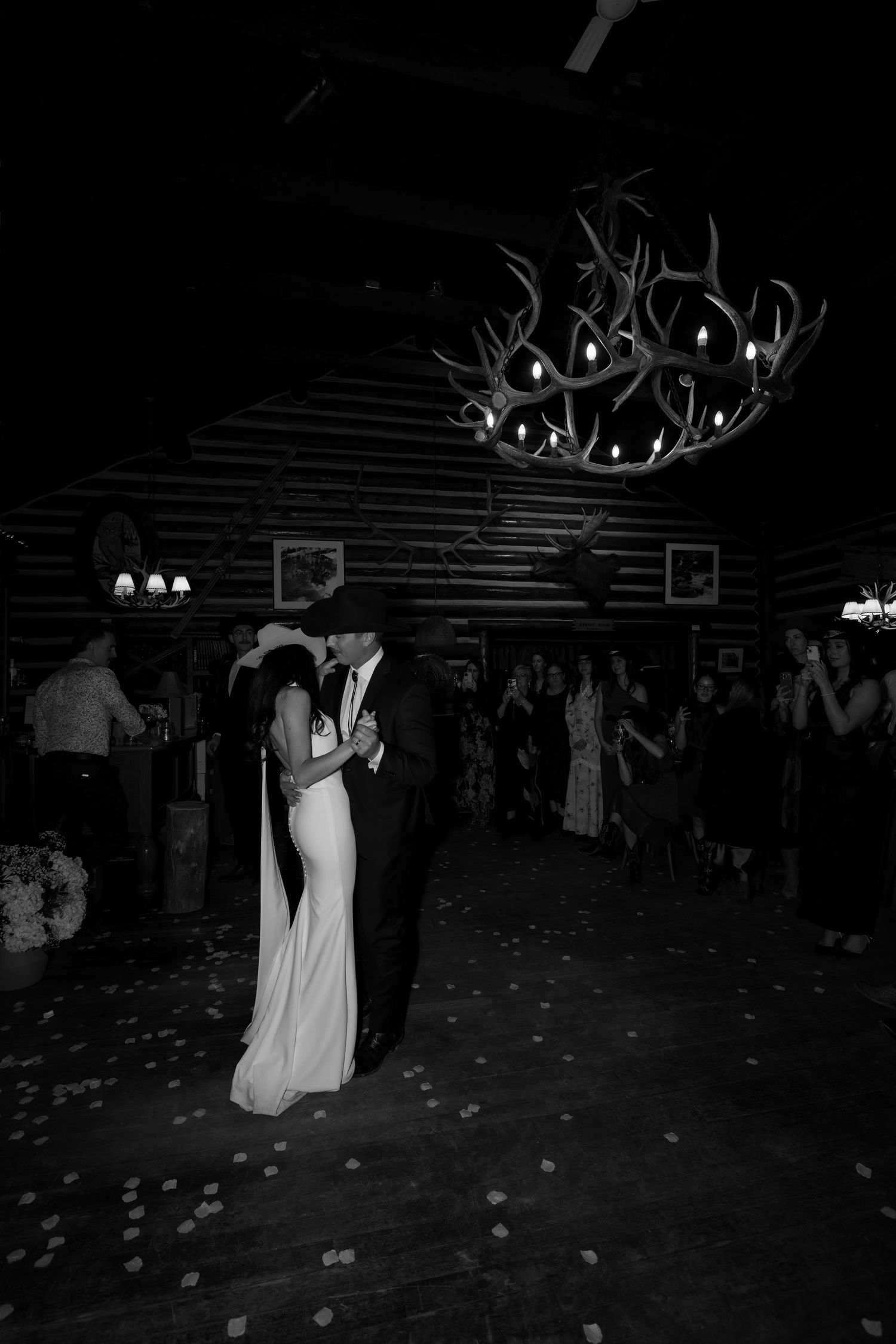 Bride and groom share first dance in black and white photo under antler chandelier in rustic log cabin venue.