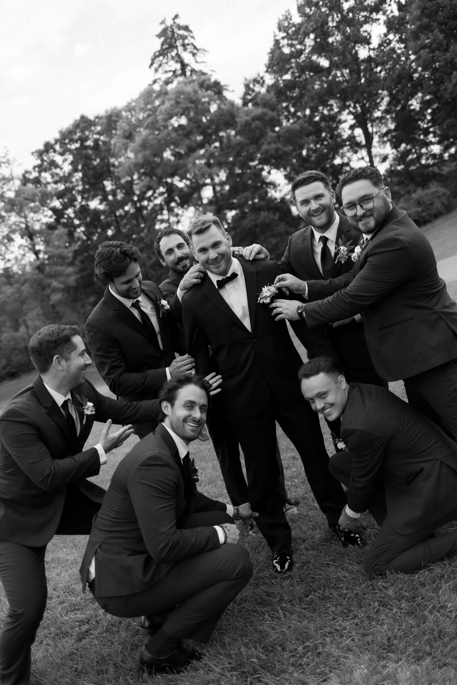 Groom laughs with groomsmen in black suits posing playfully outdoors in a fun black and white wedding photo.