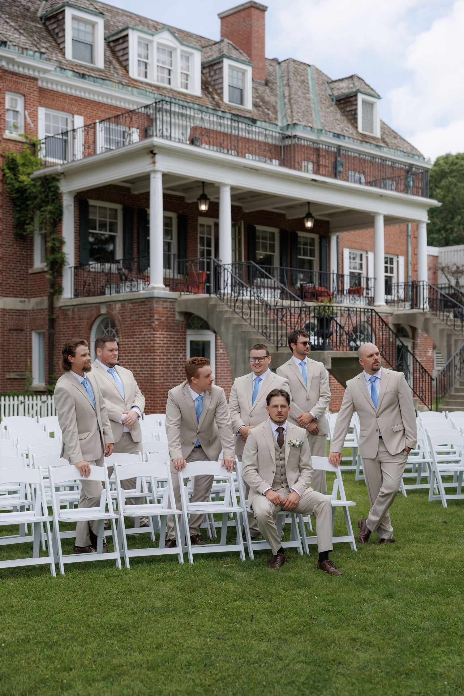 Groomsmen in tan suits with blue ties pose outdoors among white chairs at an elegant brick mansion wedding venue.
