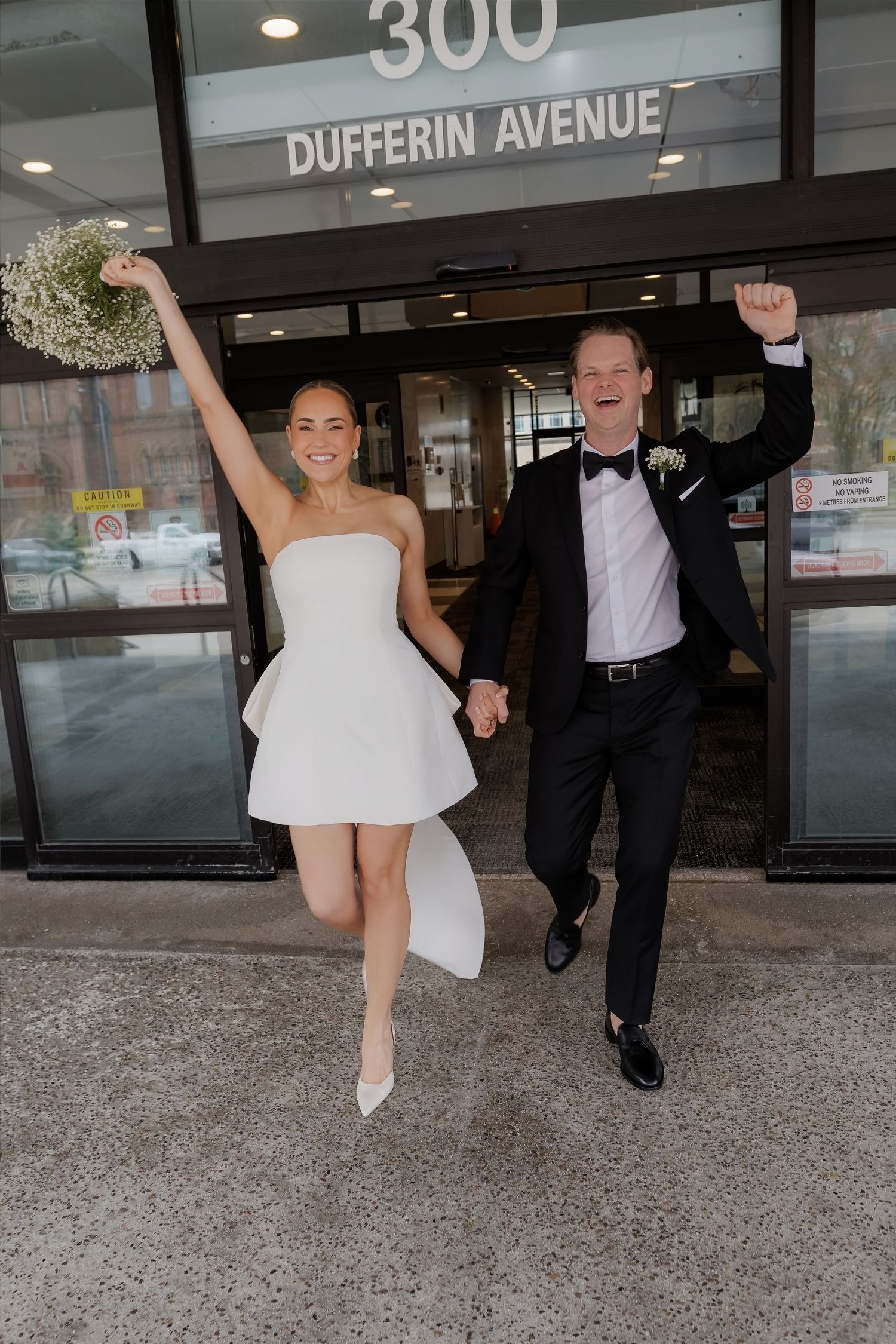 Joyful newlyweds celebrate outside 300 Dufferin Avenue, bride in mini dress holding bouquet, groom in tuxedo raising fist.