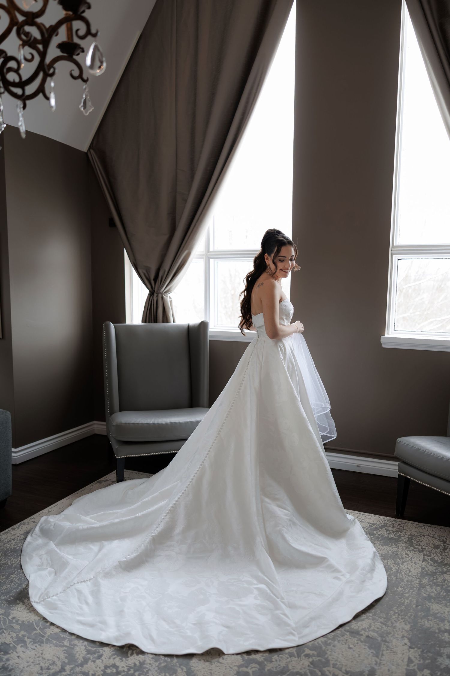 Bride in strapless ballgown with cathedral train posing by large window in elegant taupe bridal suite.