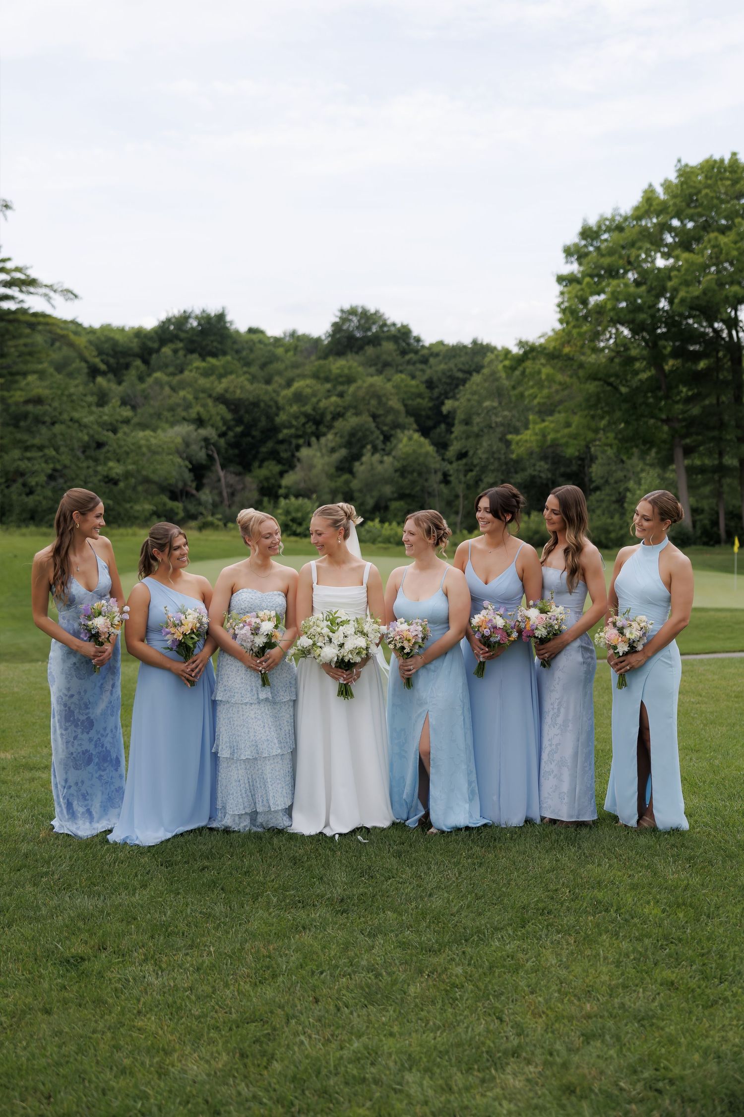 Bride with six bridesmaids in light blue gowns holding bouquets on a green lawn outdoors.