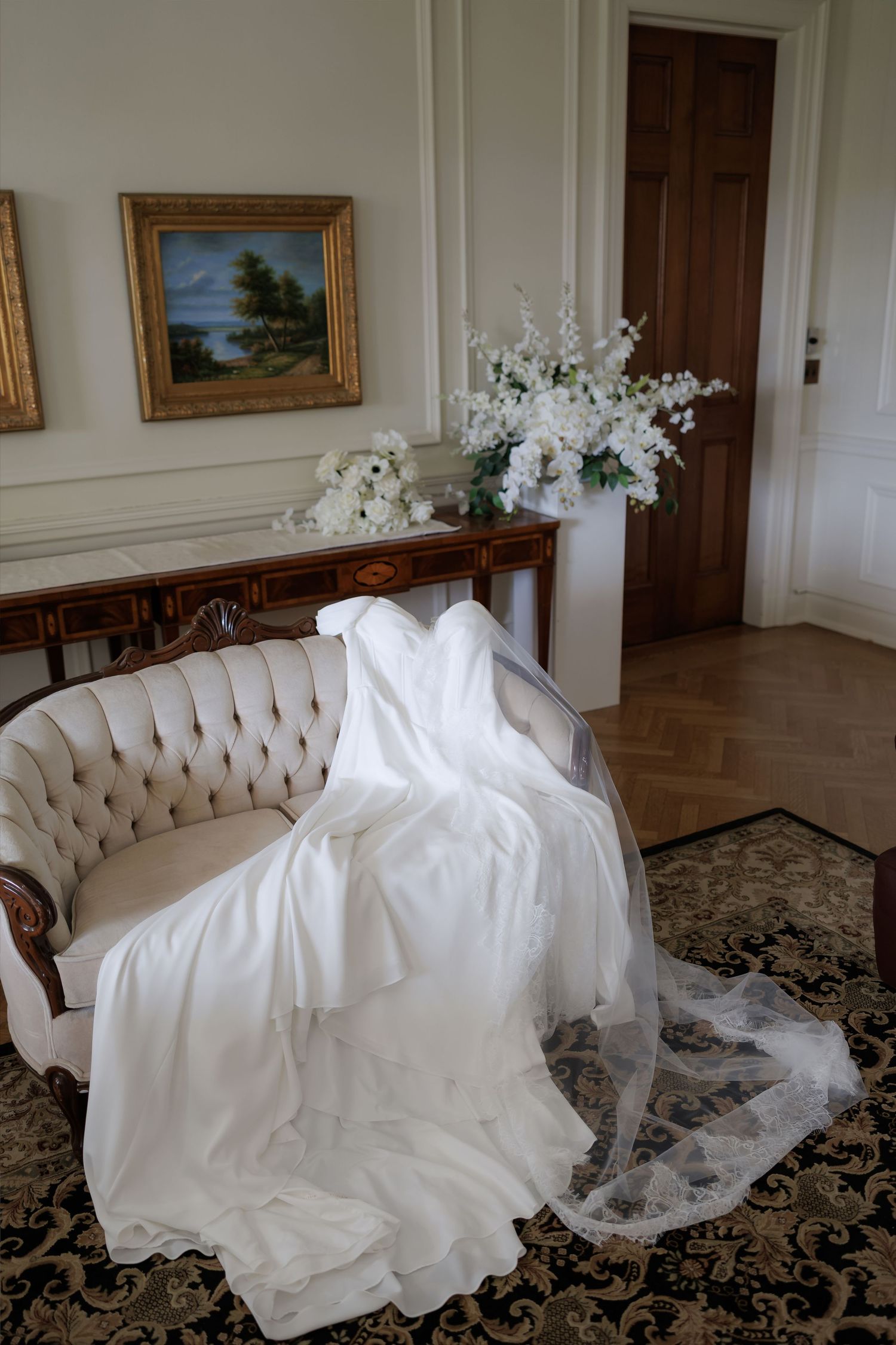 White wedding dress and veil draped over a tufted cream sofa in an elegant room with floral arrangements and gold-framed art.