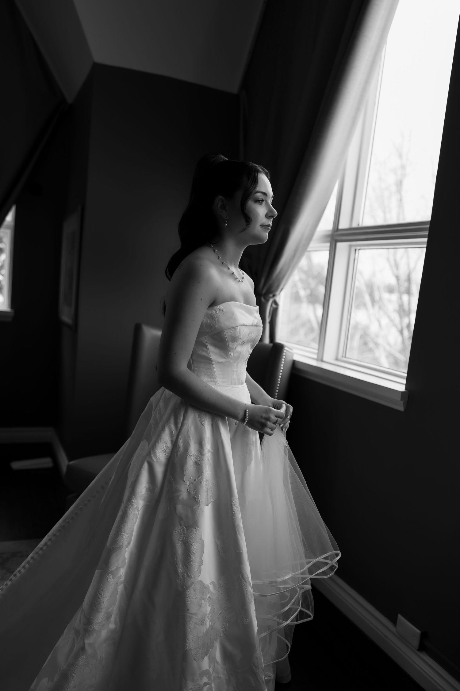 Black and white photo of a bride in a strapless ball gown standing by a window, gazing outside on her wedding day.