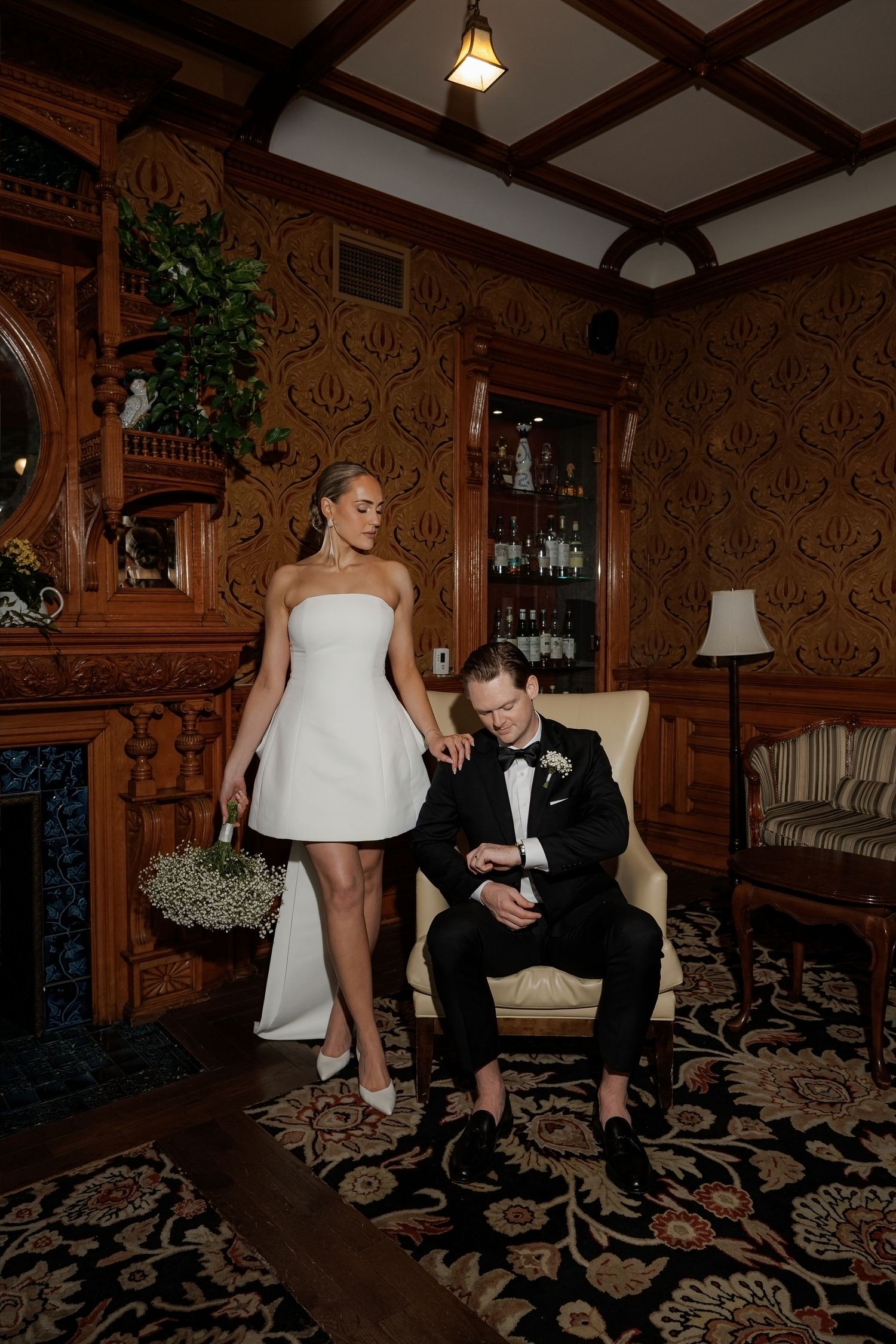 Bride in white mini dress and groom in tuxedo pose in elegant wood-paneled room during wedding portraits.