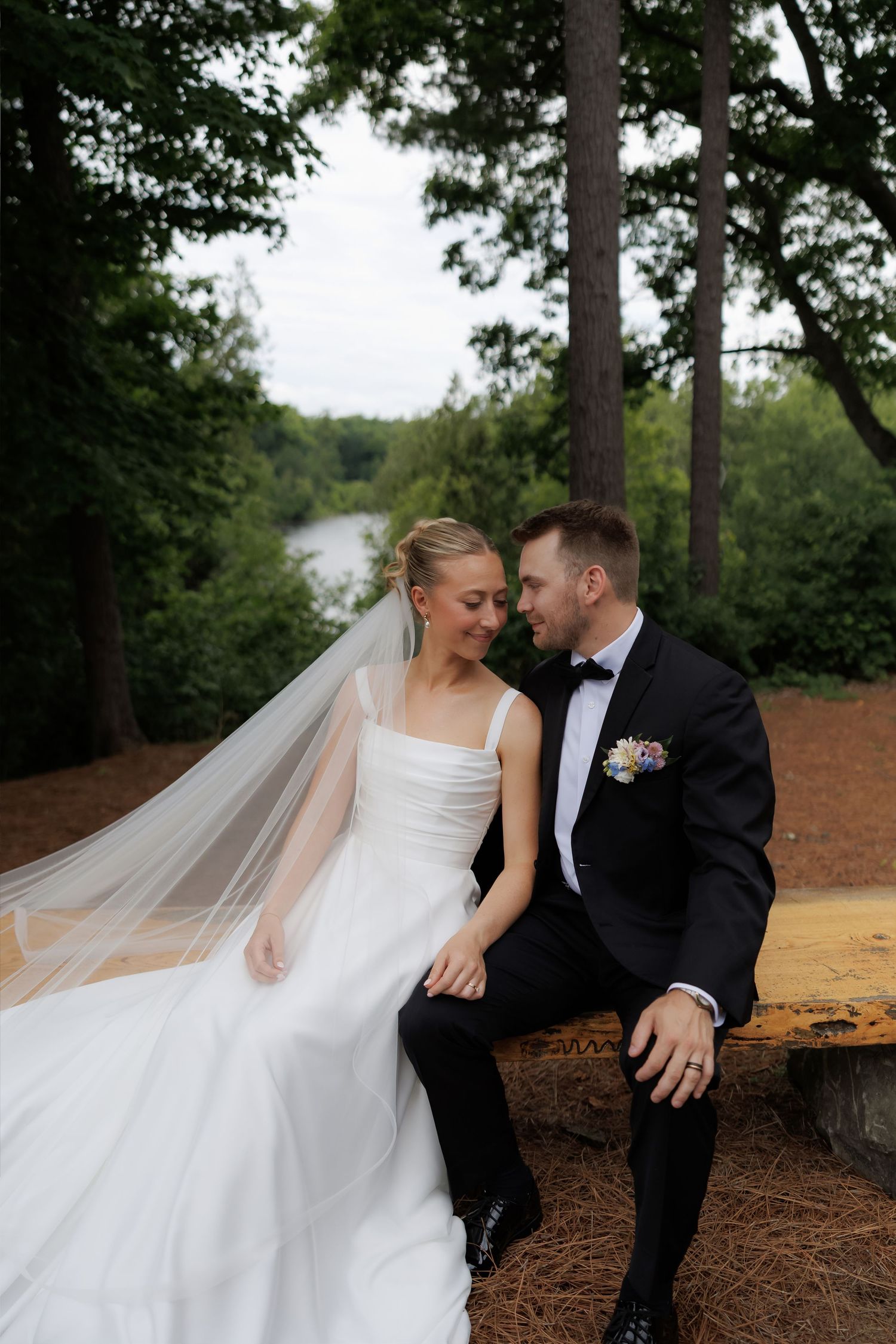 Bride and groom sitting on a wooden bench in a lush forest setting near a lake, sharing an intimate moment.