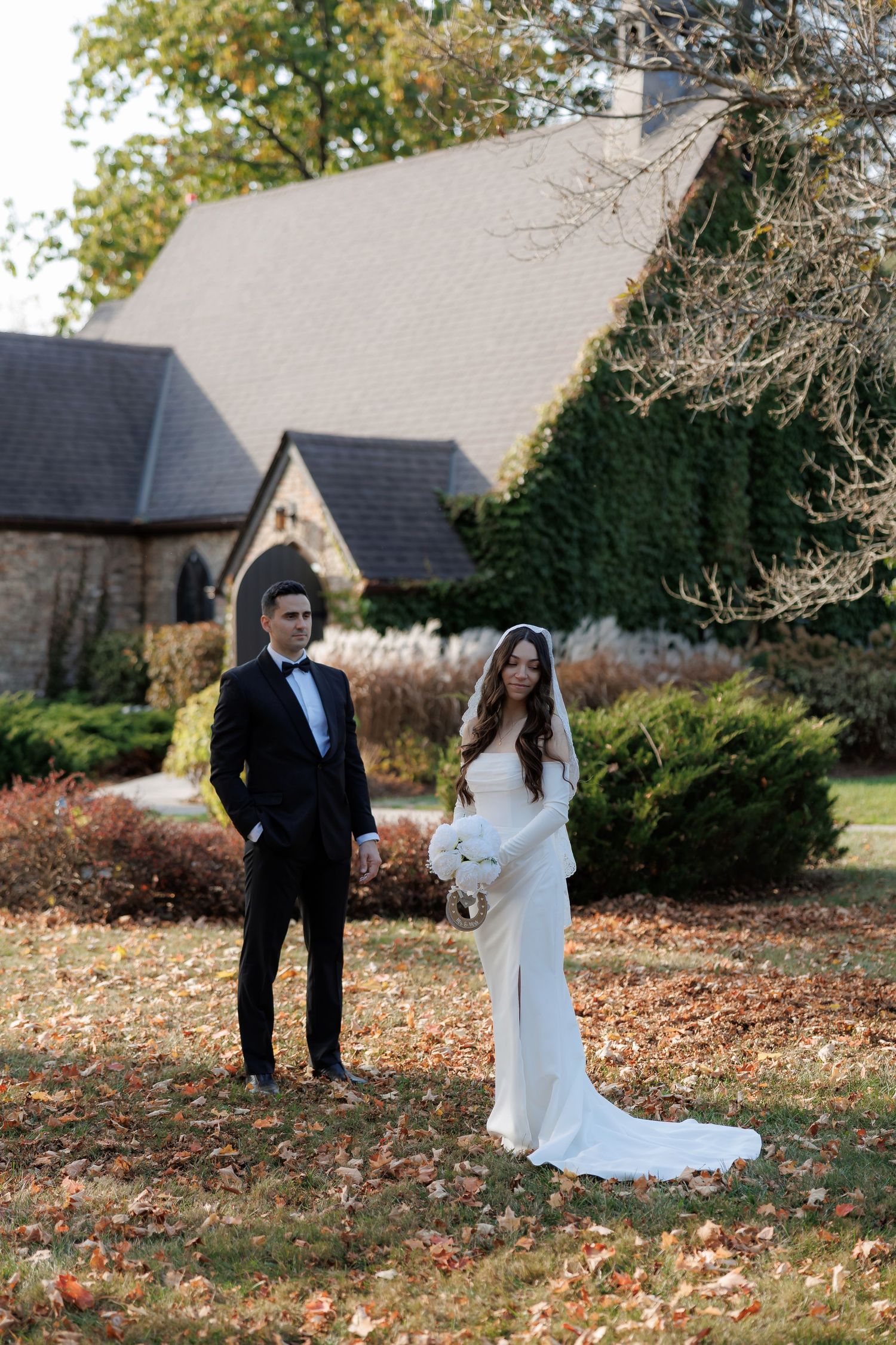 Wedding couple poses in a garden with autumn leaves and buildings in background.