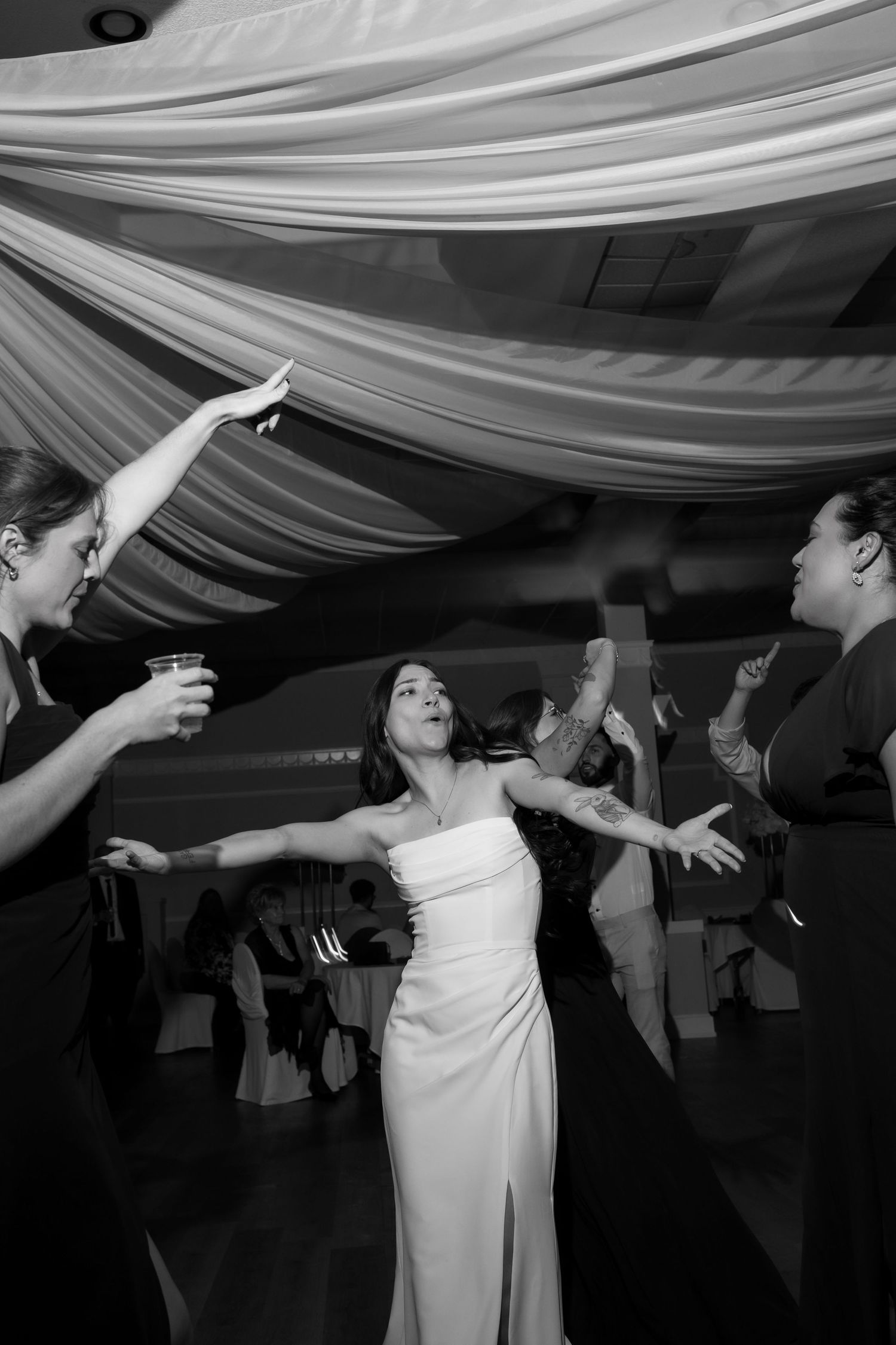 Wedding party members dance with enthusiasm under elegant white ceiling draping.