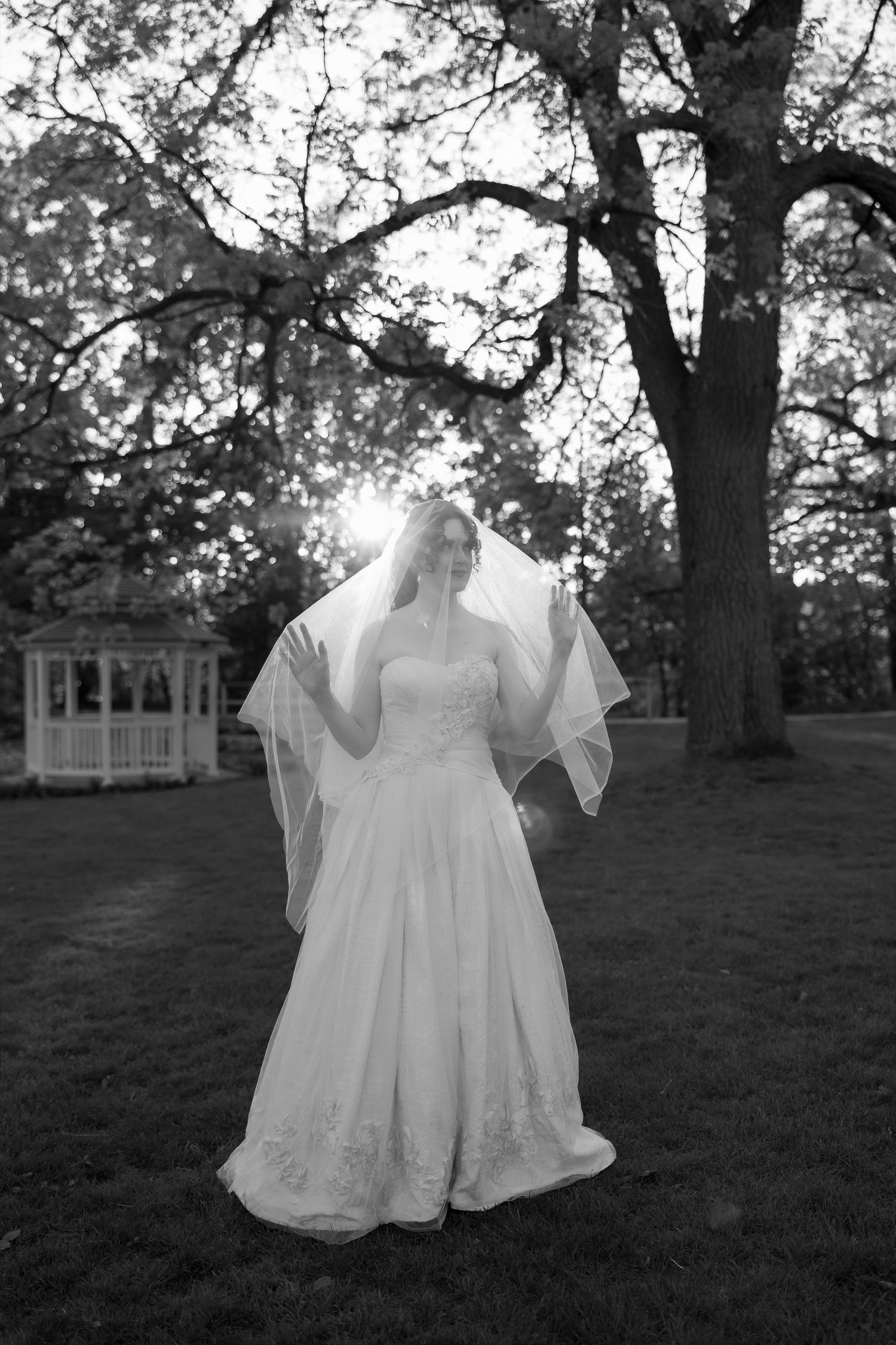 Bride in white gown and veil stands outdoors near gazebo and large tree in black and white wedding portrait.