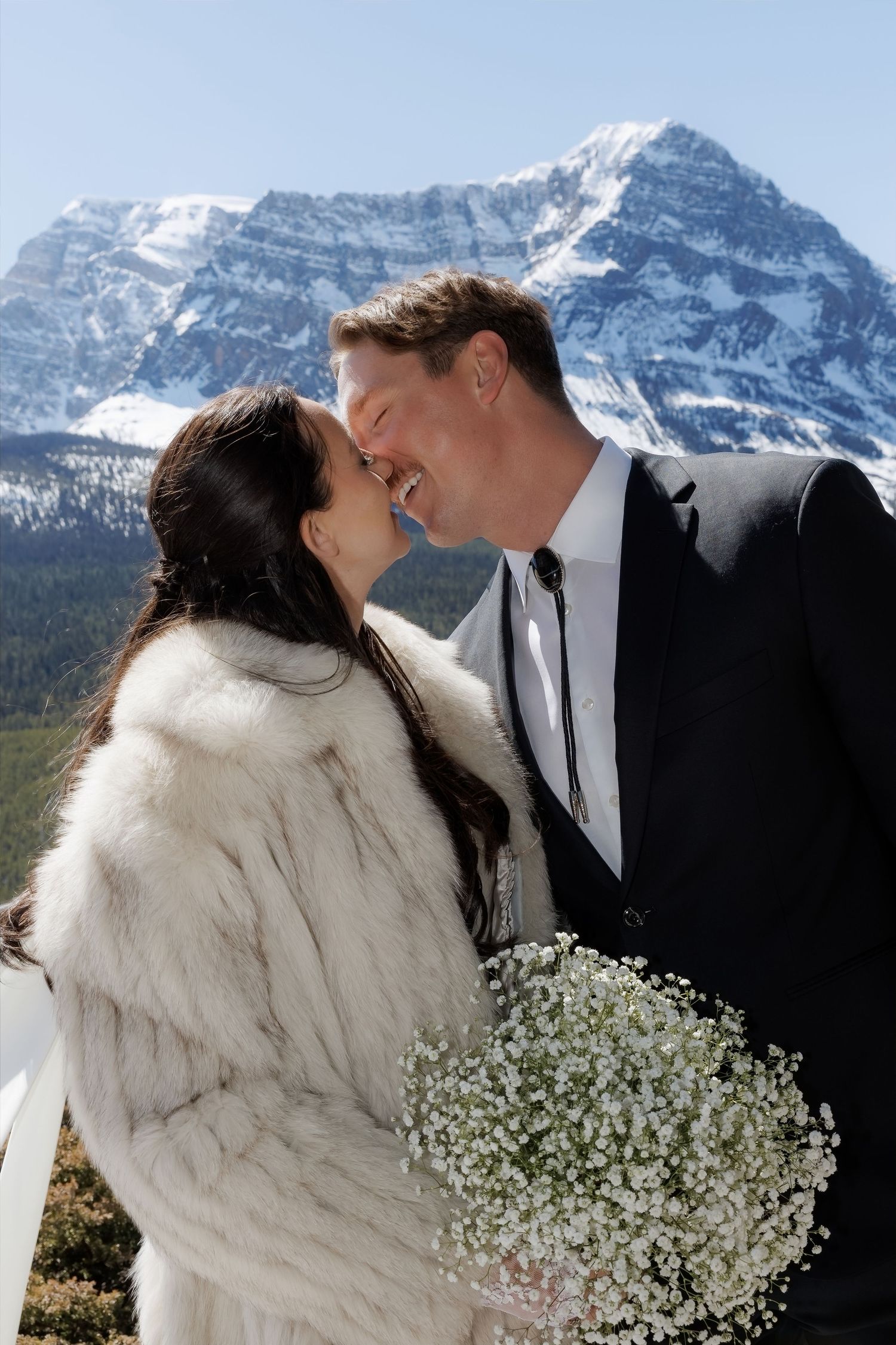 Bride and groom share a kiss with snow-capped mountain backdrop, bride wearing white fur coat holding baby's breath bouquet.