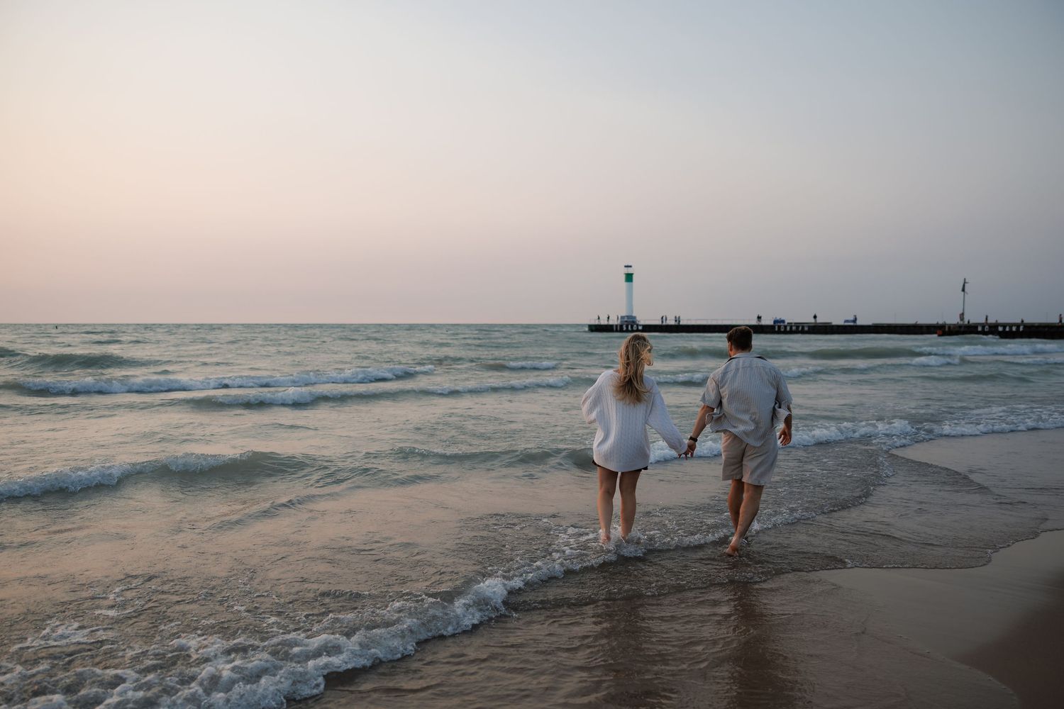 Couple holding hands walking along sandy beach at sunset near a pier with lighthouse.