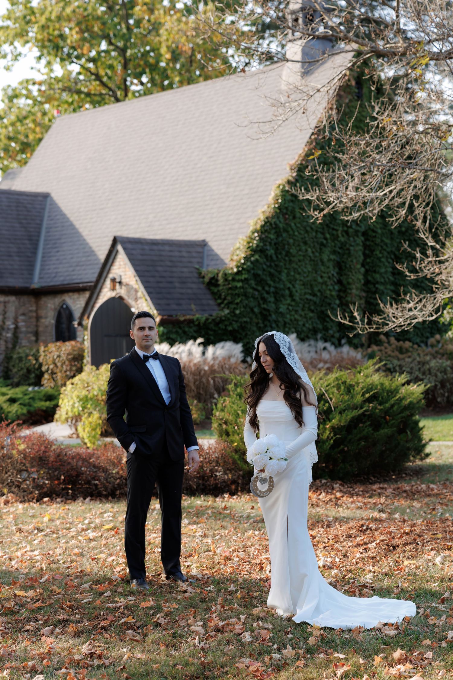 Fall wedding portrait with bride and groom standing in front of stone buildings surrounded by autumn leaves.