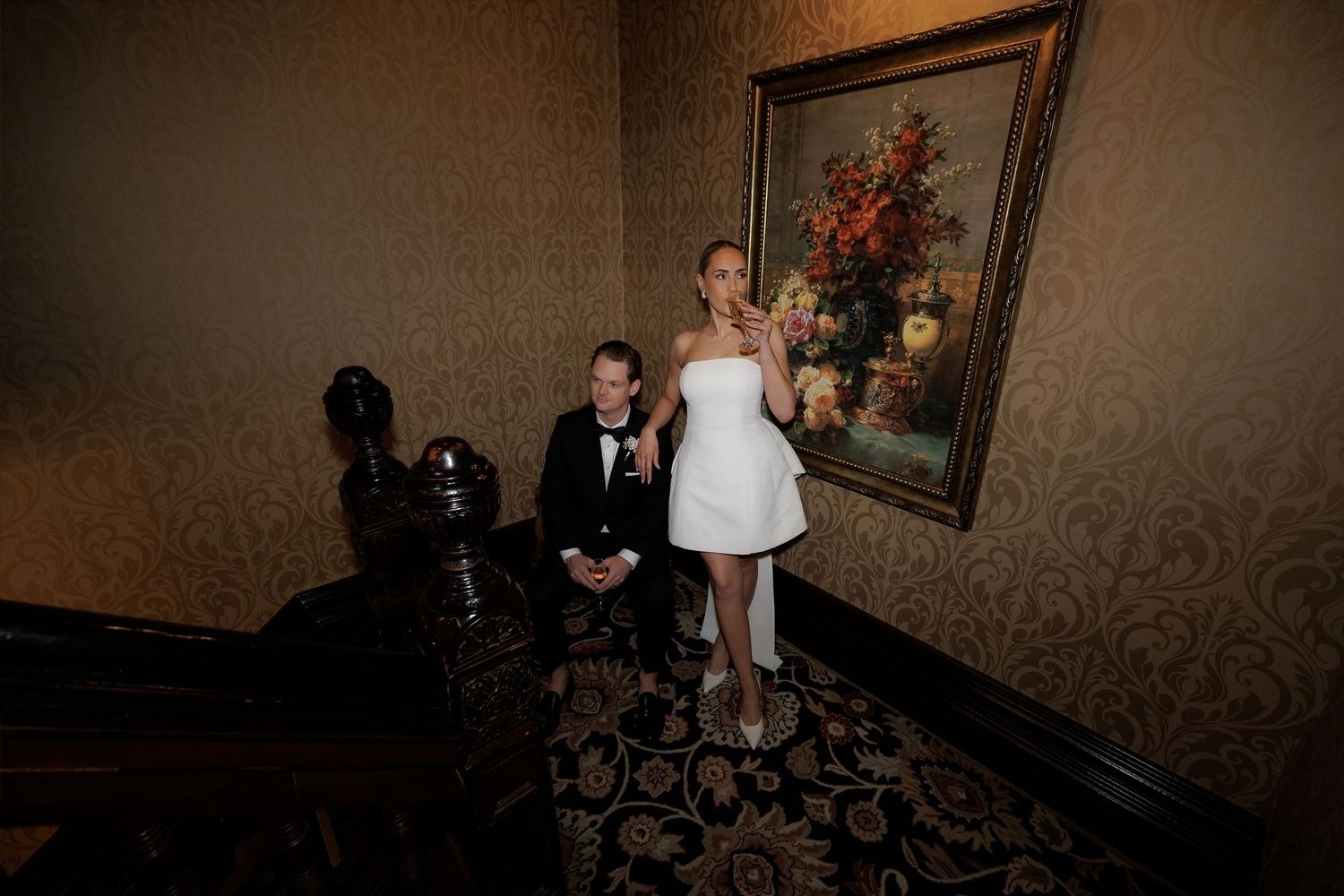 Elegant bride in white mini dress and groom in tuxedo posing in ornate room with floral painting.