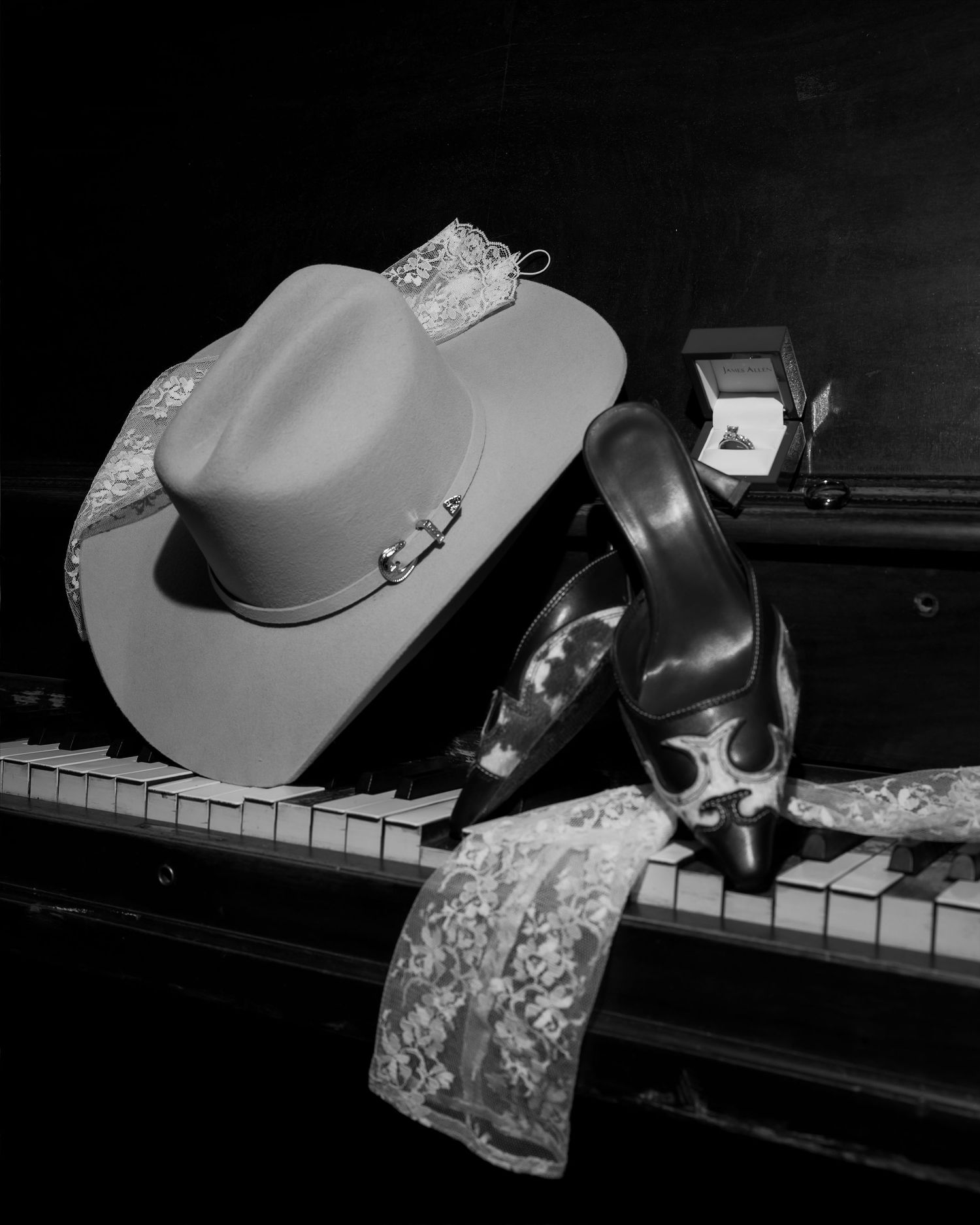 Black and white photo of cowboy hat, heels, lace, and ring box on piano keys.
