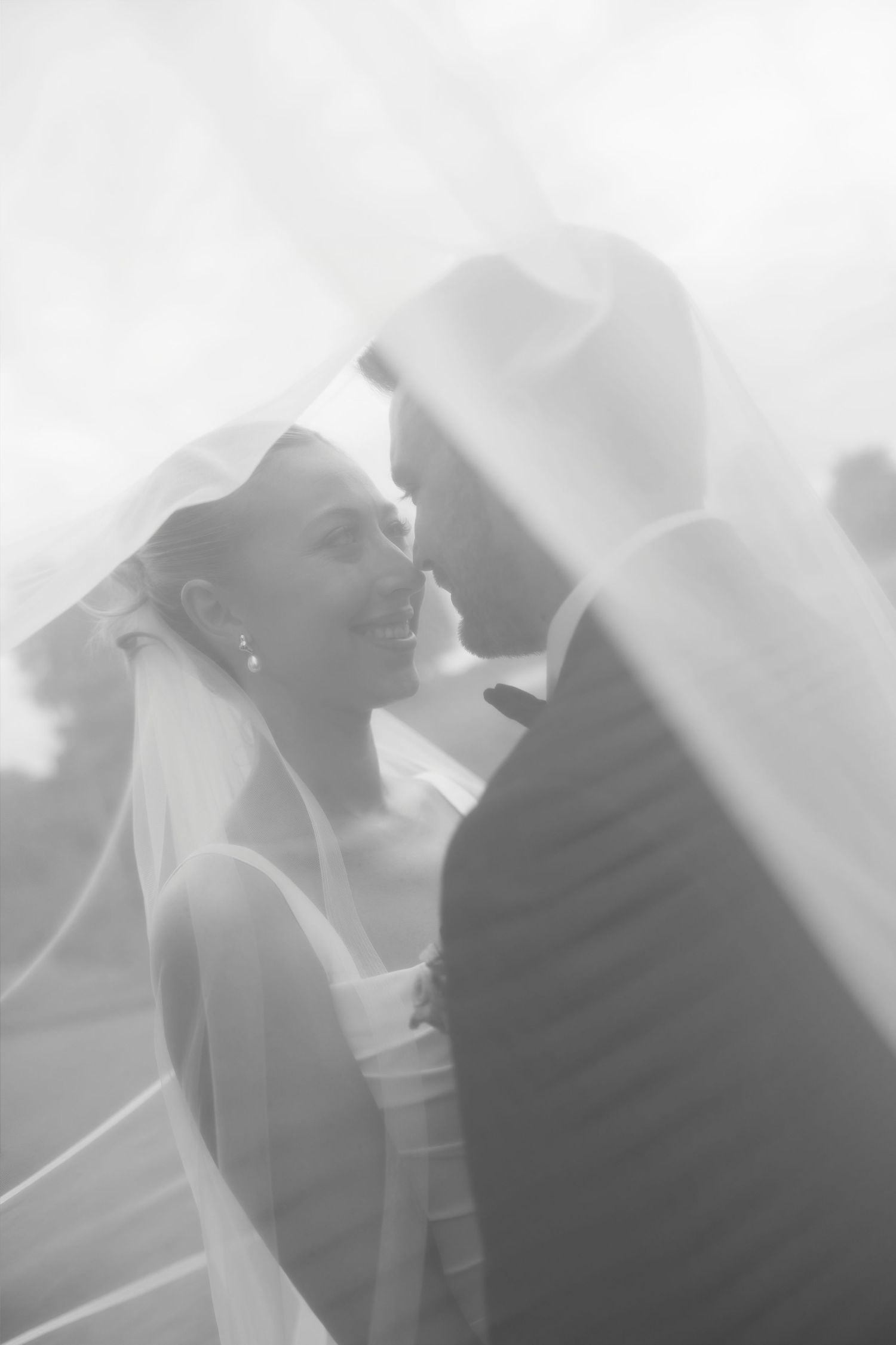 Black and white photo of smiling bride and groom sharing romantic moment under flowing wedding veil.