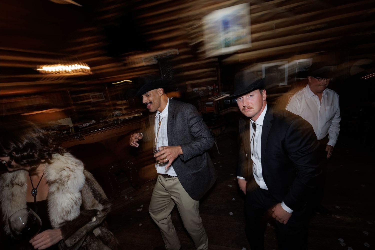 Men in cowboy hats and suits dance energetically at a rustic western-themed wedding reception.