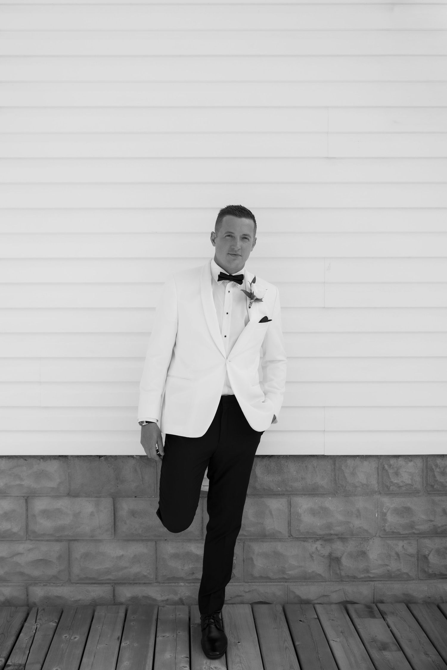 A person in a white tuxedo jacket and black pants poses casually against a white wall.