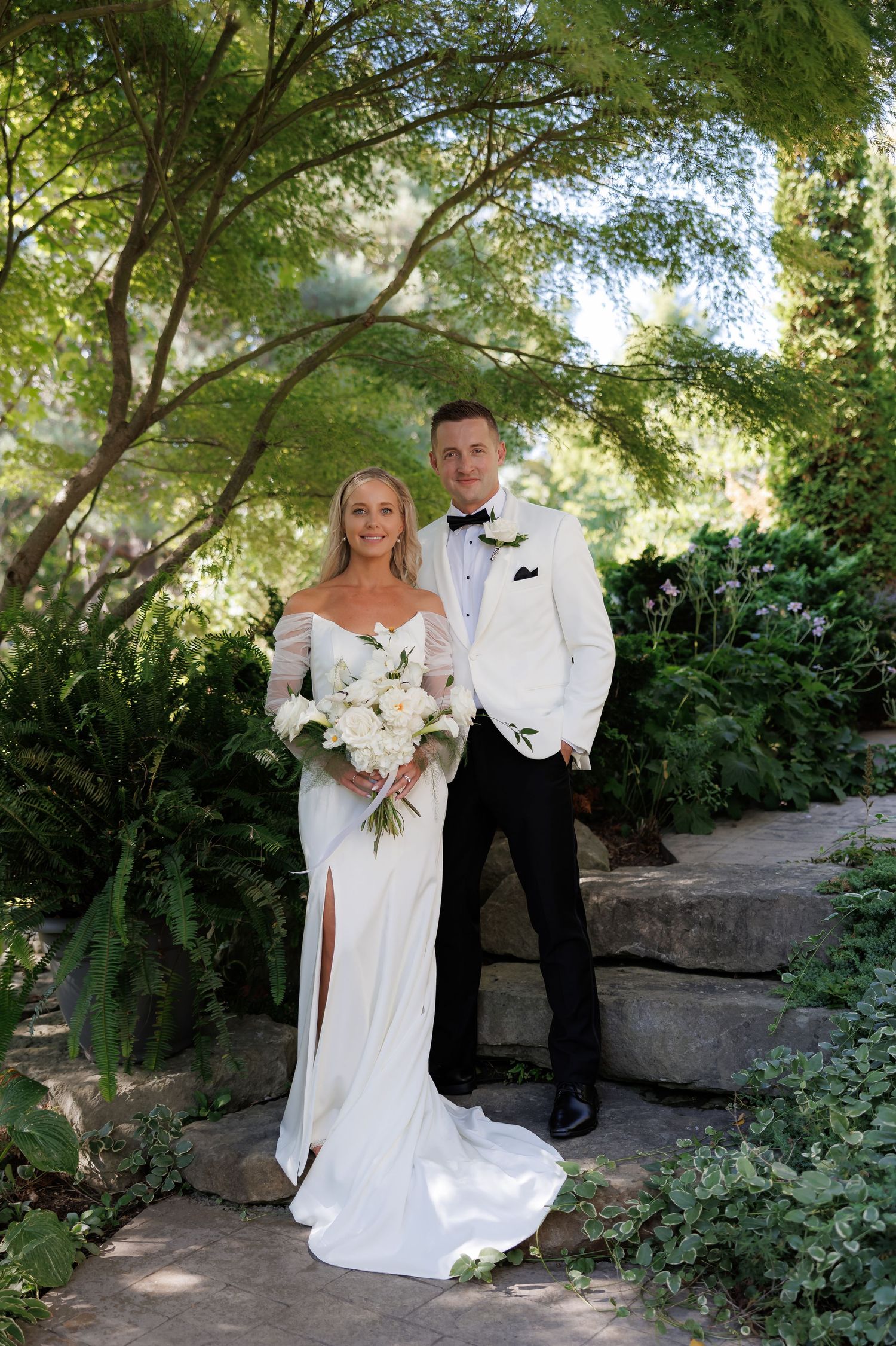 Newlyweds share a romantic moment in a sunlit garden pathway surrounded by greenery.