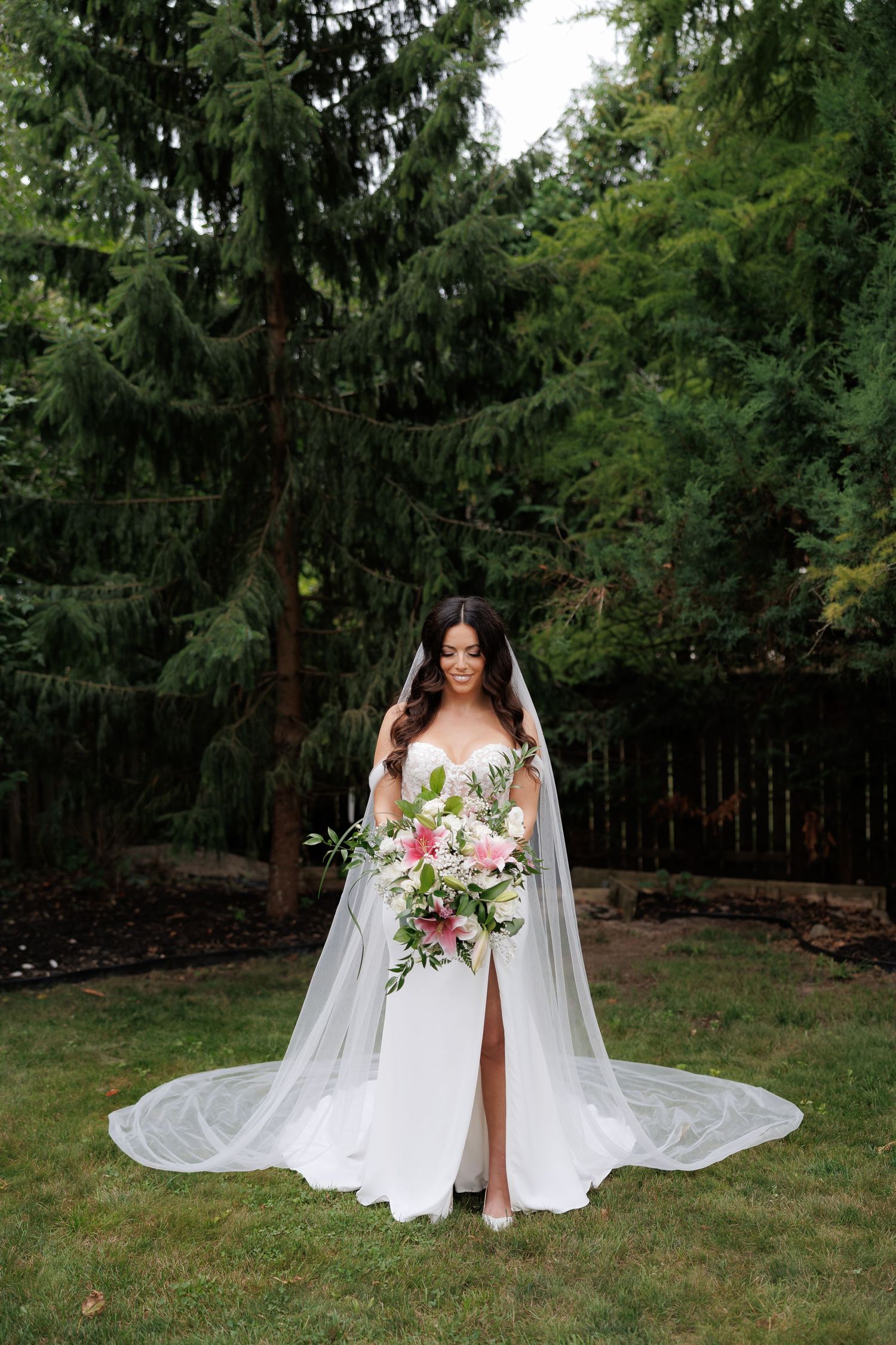 Bride in white gown with flowing veil poses with bouquet in garden setting.