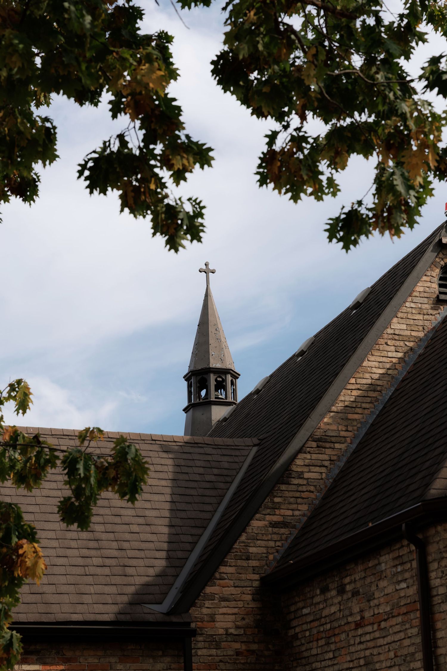 Church roofline with cross and steeple against cloudy sky framed by autumn leaves.