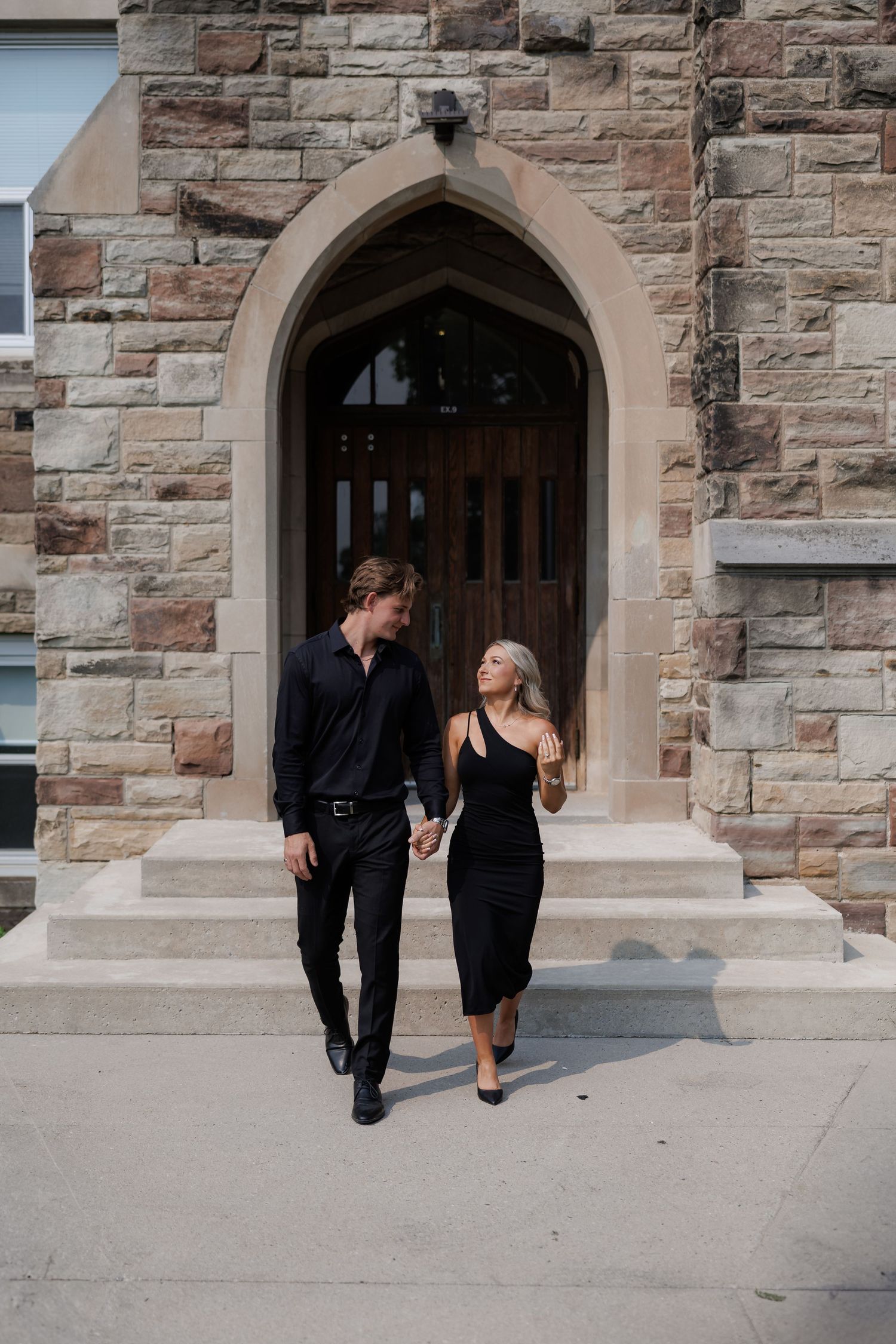 Stylish couple in black outfits walking through a Gothic stone archway.