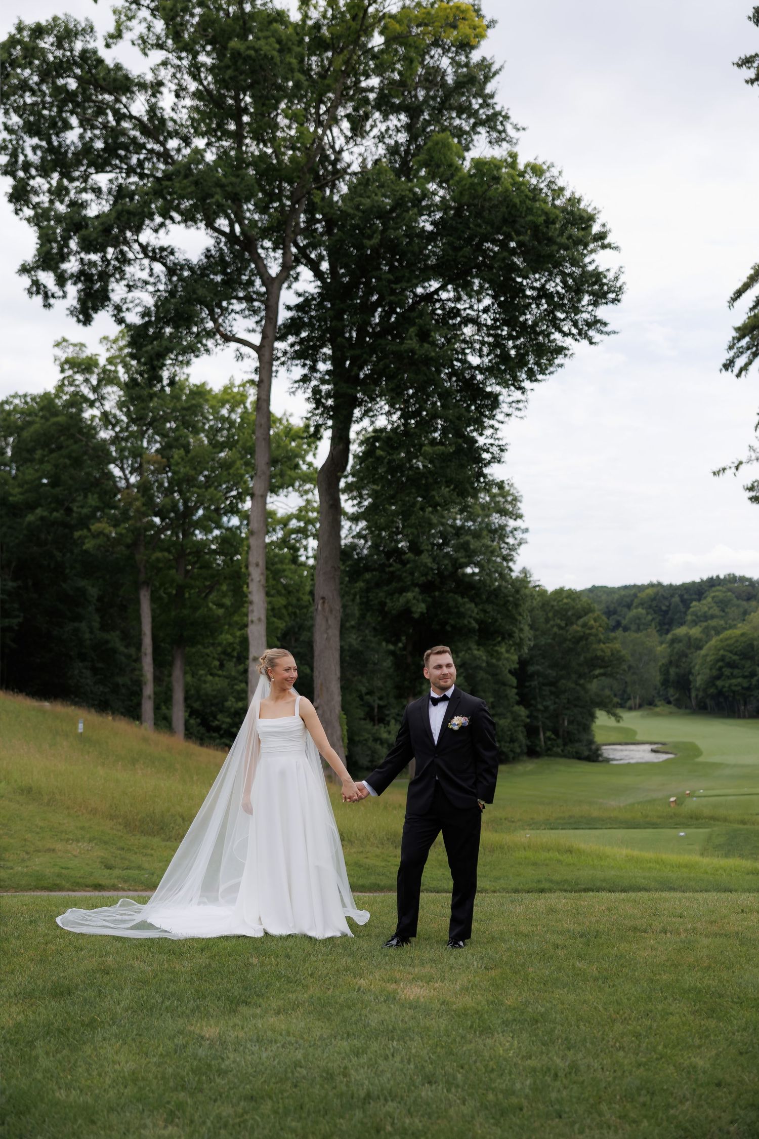 Bride and groom holding hands on lush green golf course surrounded by tall trees on wedding day.
