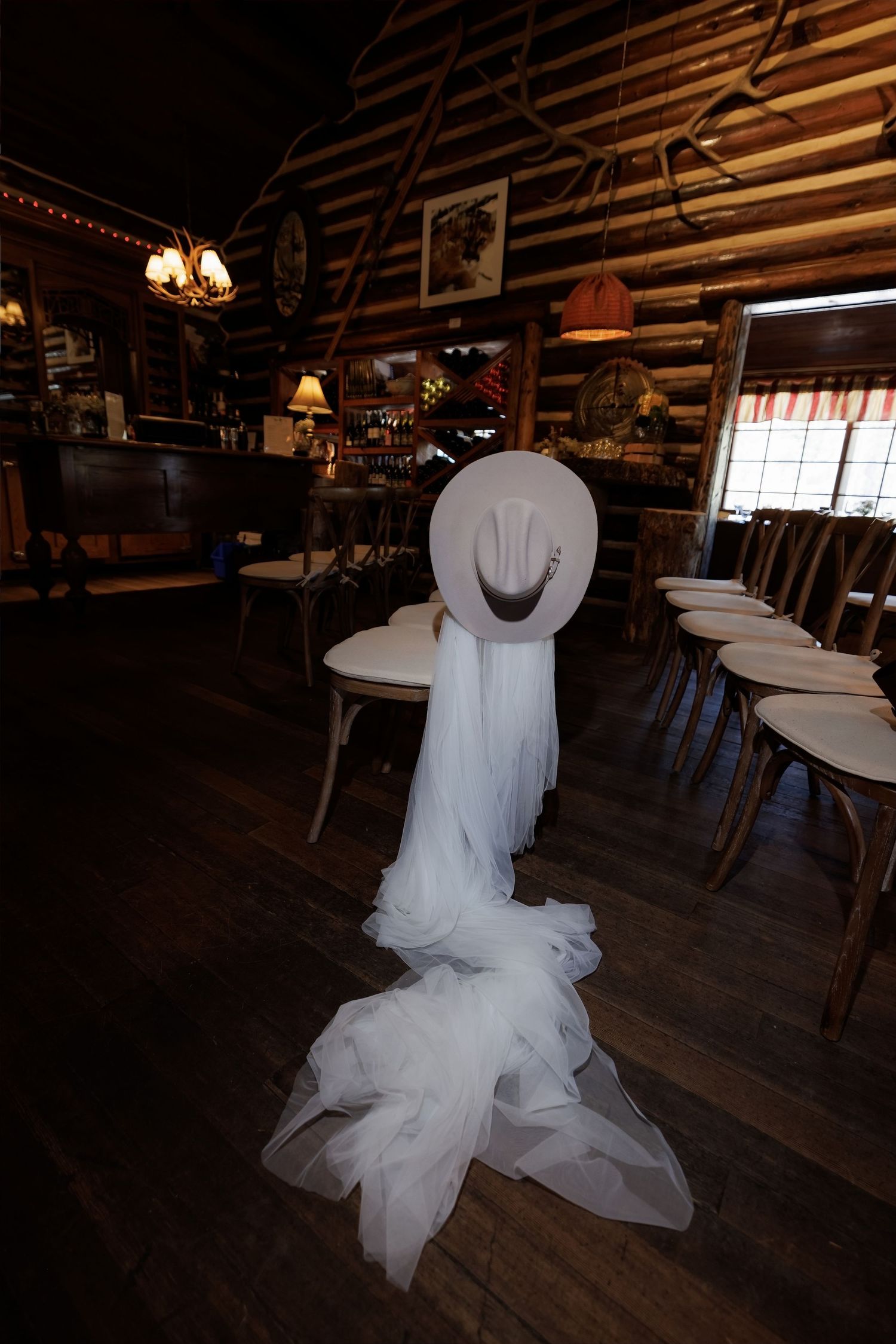 White cowboy hat and flowing veil draped over chair in rustic log cabin wedding venue with wood interior.