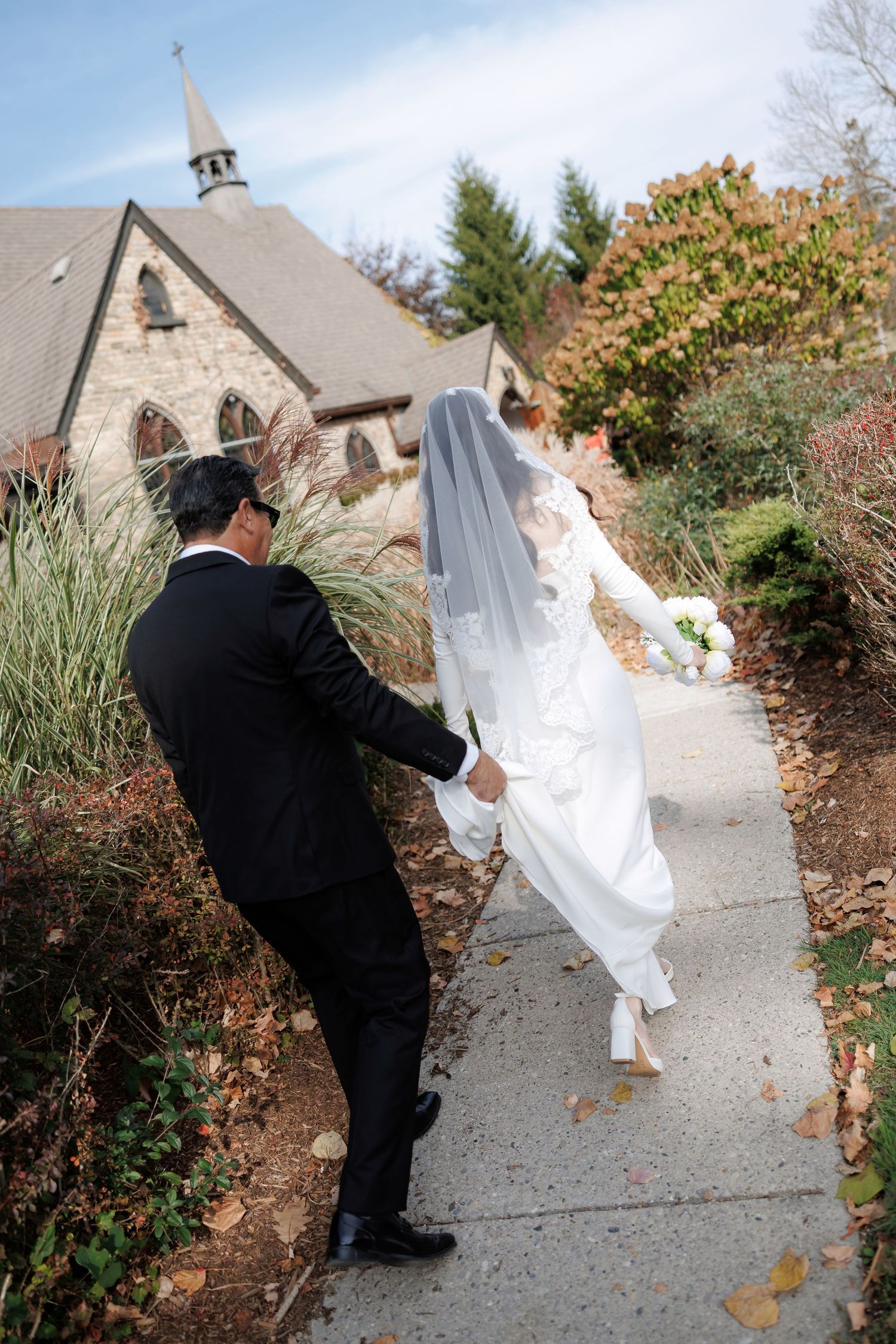 Wedding couple walks along church pathway surrounded by autumn foliage.