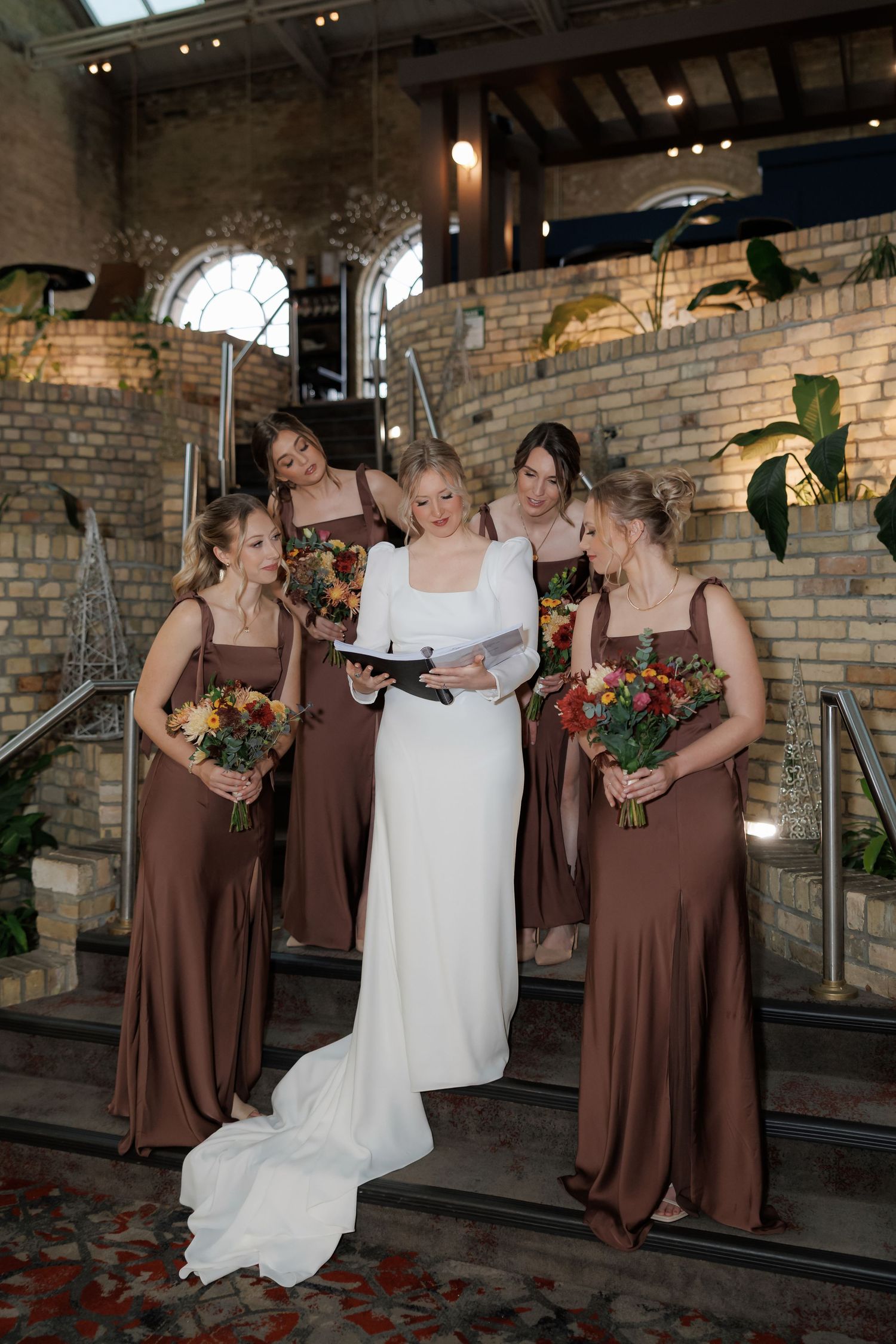 Wedding party in brown attire shares candid moment against rustic brick backdrop.