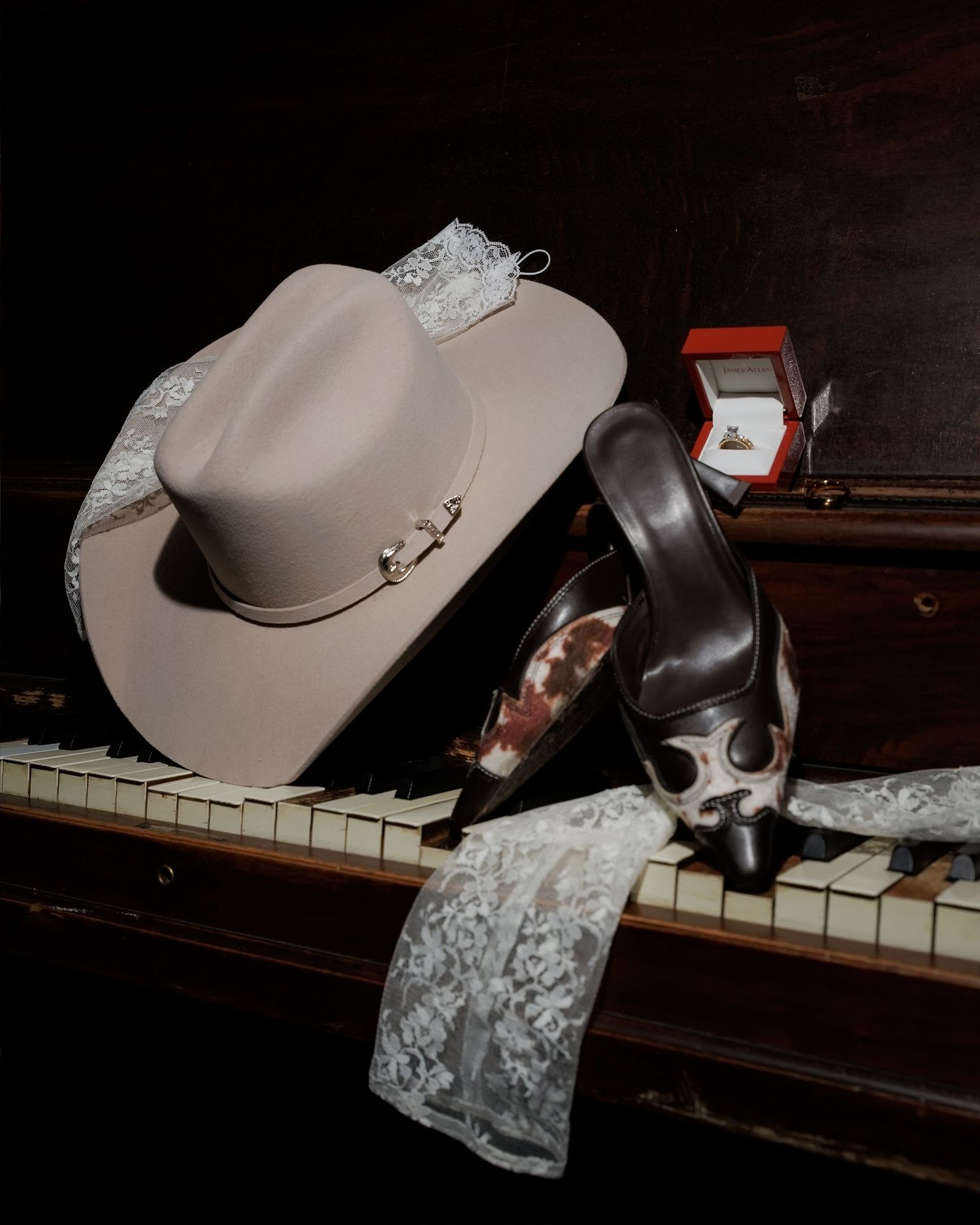 Elegant wedding accessories including a tan cowboy hat, heels, lace, and a ring box displayed on piano keys.