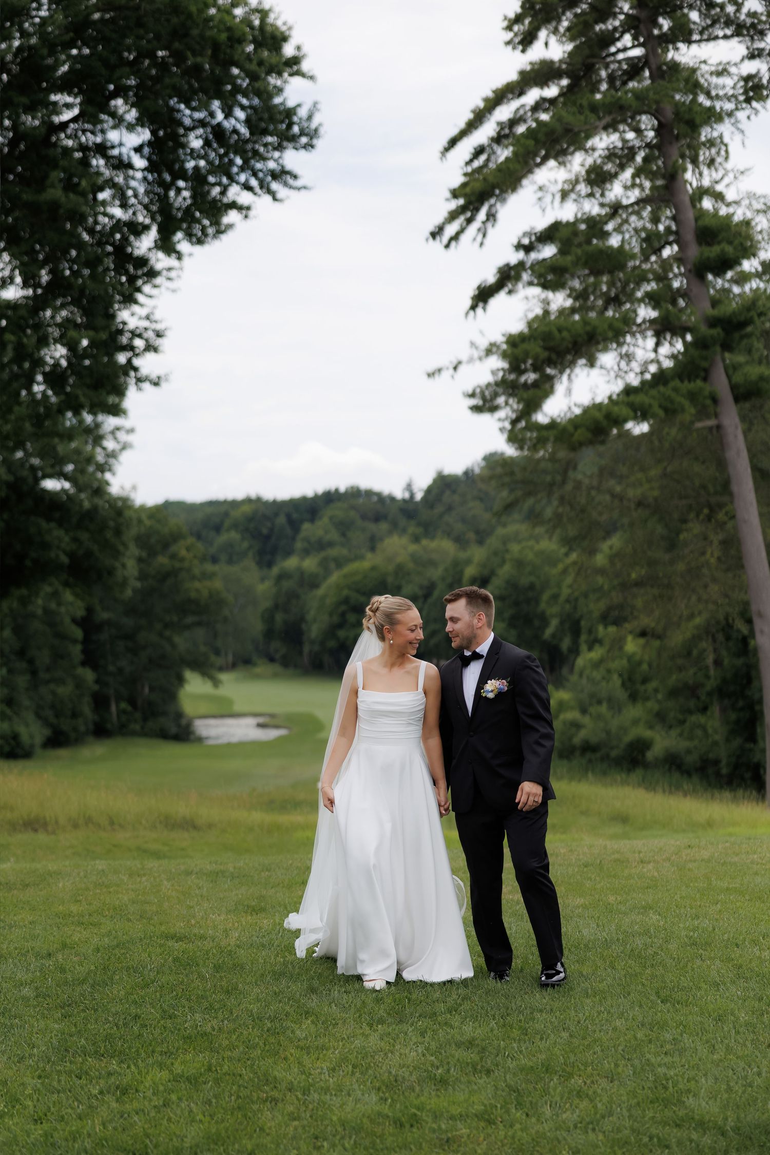 Bride and groom walk hand in hand on lush green golf course surrounded by tall trees on their wedding day.