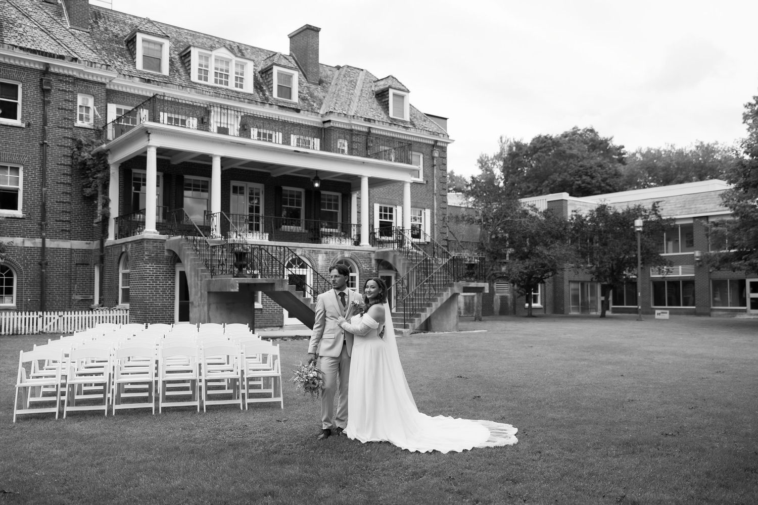 Black and white photo of bride and groom posing on lawn before elegant brick mansion with wedding chairs arranged outside.
