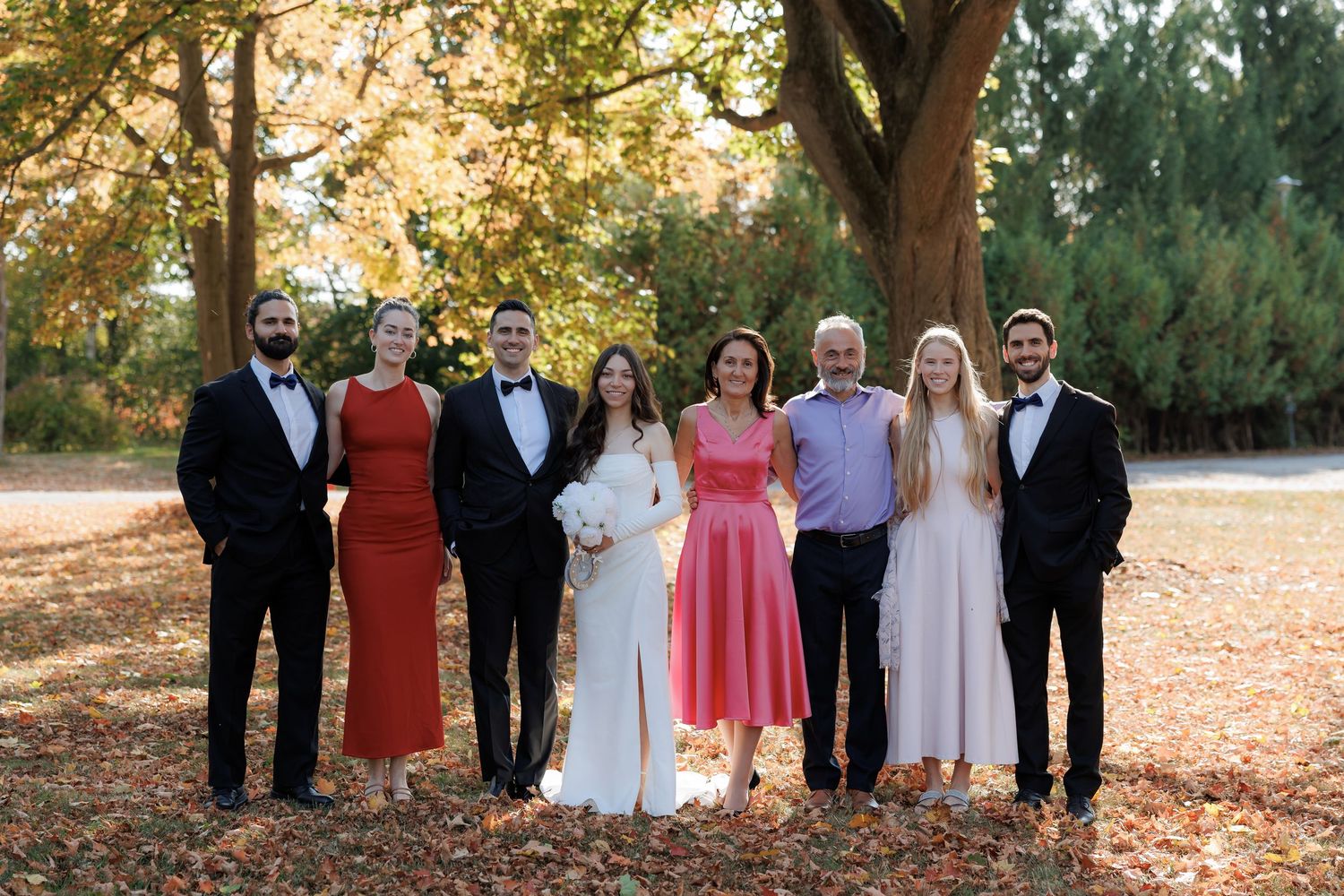 Wedding party in formal attire poses together under autumn trees.