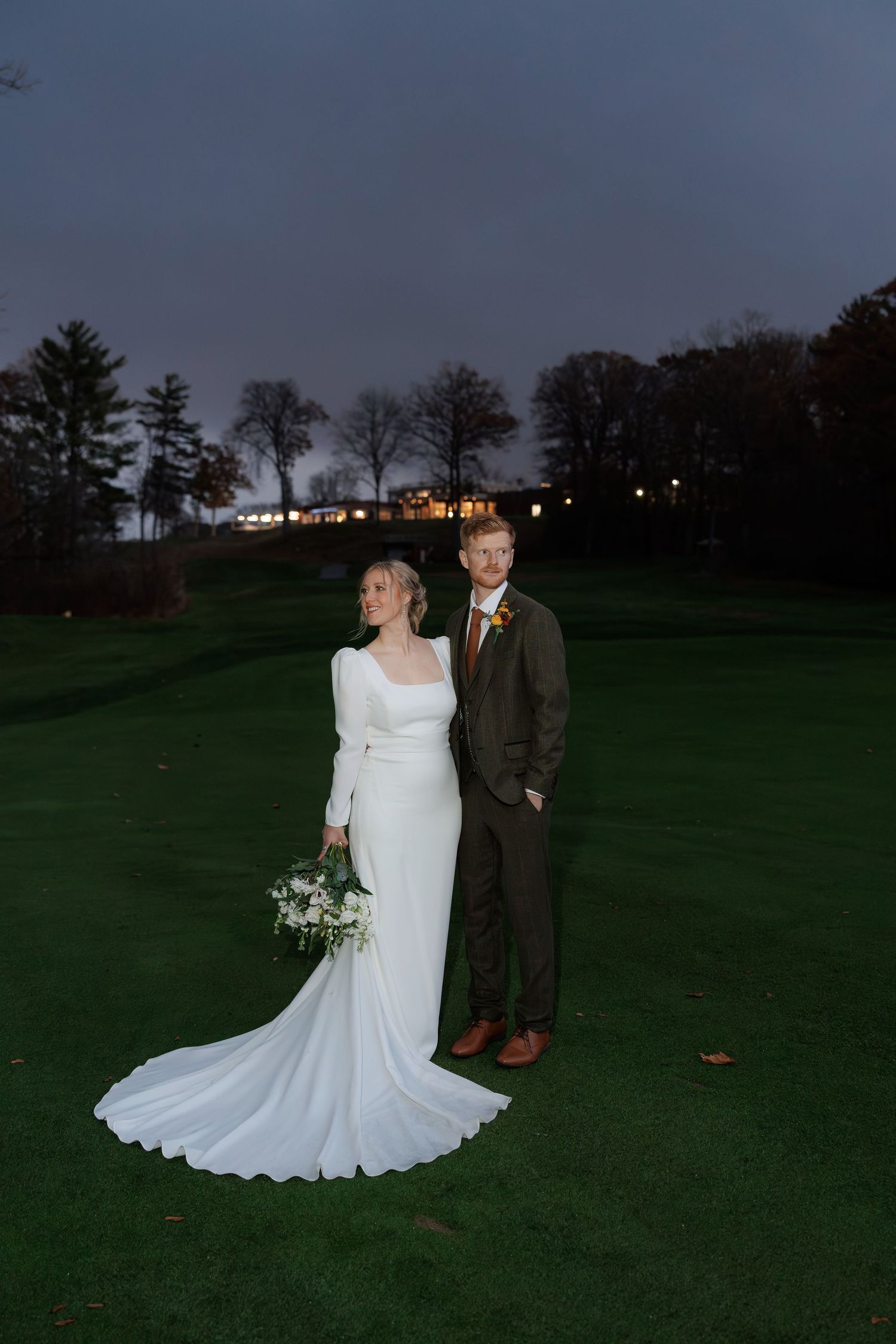 Wedding couple poses on a golf course at twilight with distant lights creating a romantic ambiance.