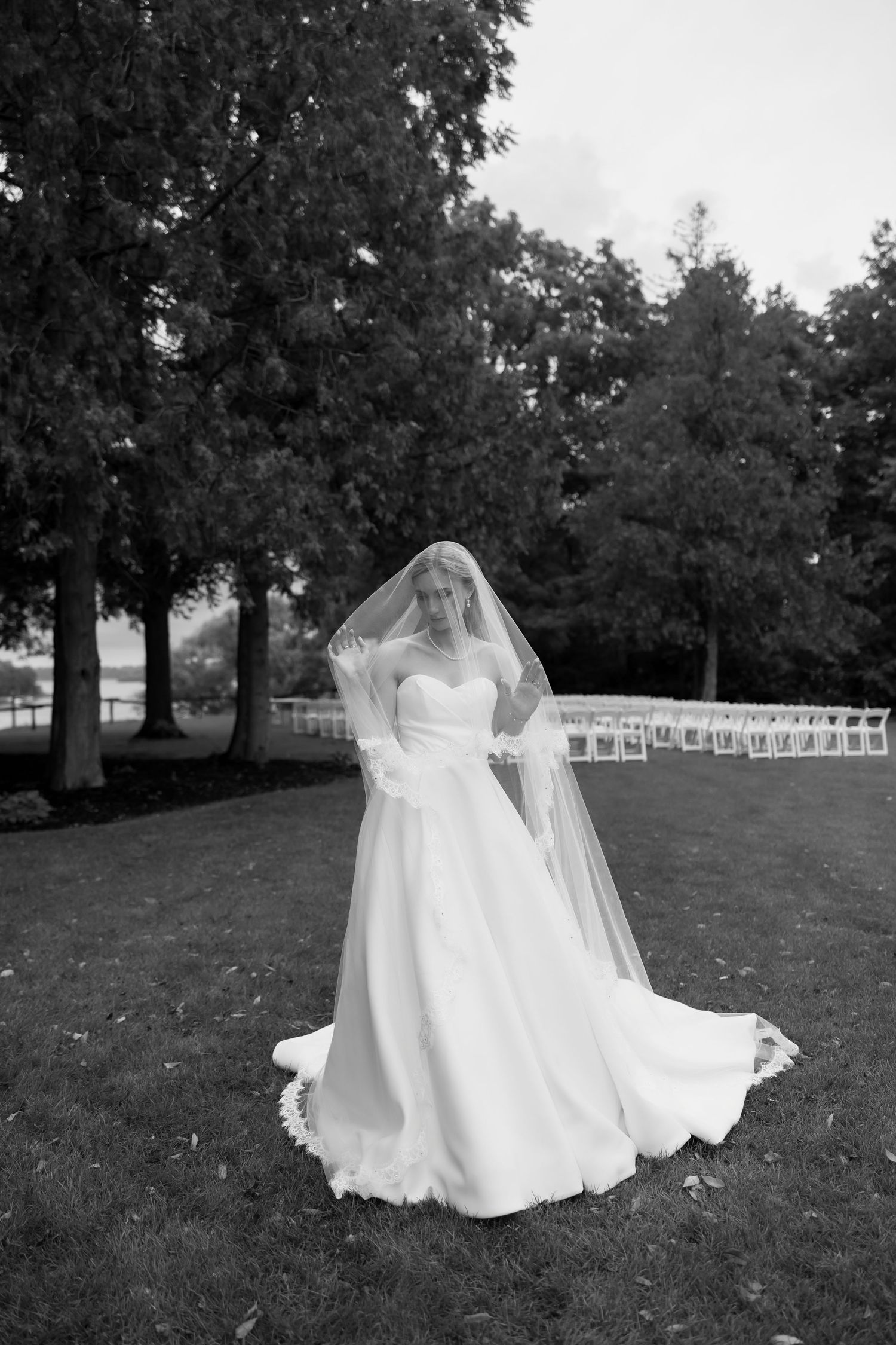 Bride in strapless ball gown with flowing veil stands on lush lawn surrounded by trees at outdoor wedding venue.