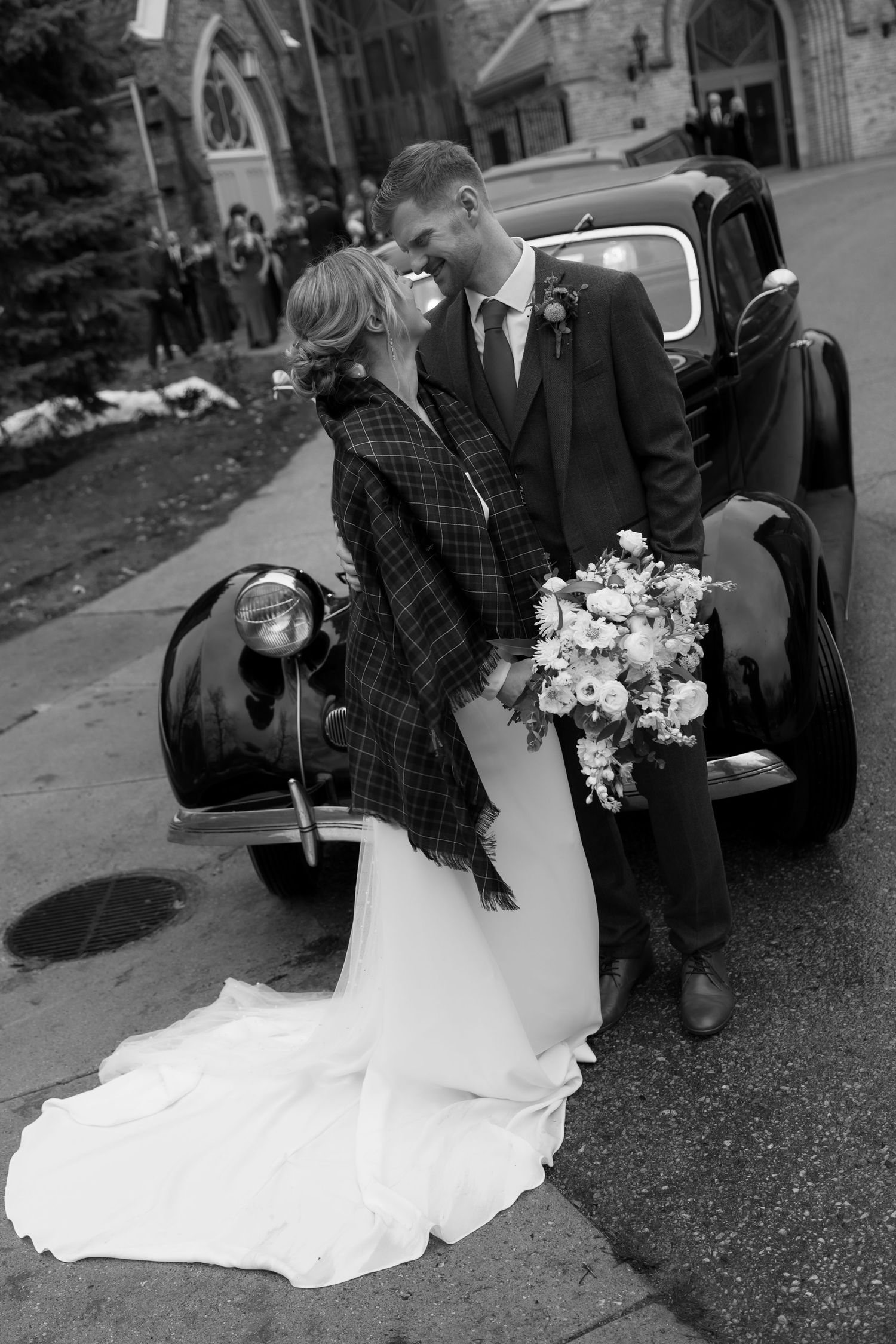 A romantic black and white wedding photo with a couple kissing next to a vintage car outside a church.