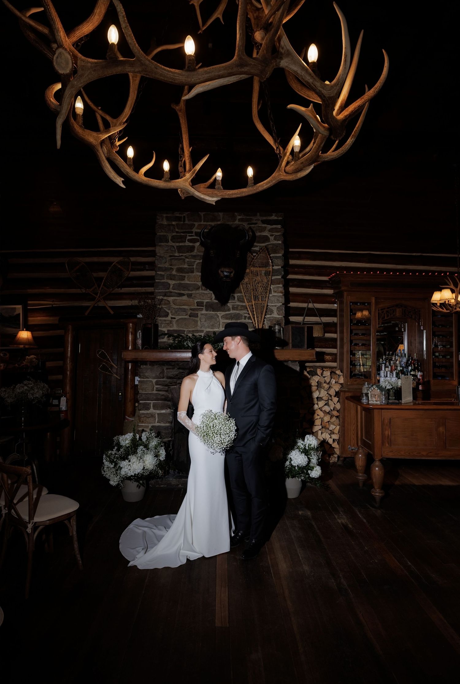 Bride and groom pose under rustic antler chandelier by stone fireplace in elegant log cabin wedding venue.