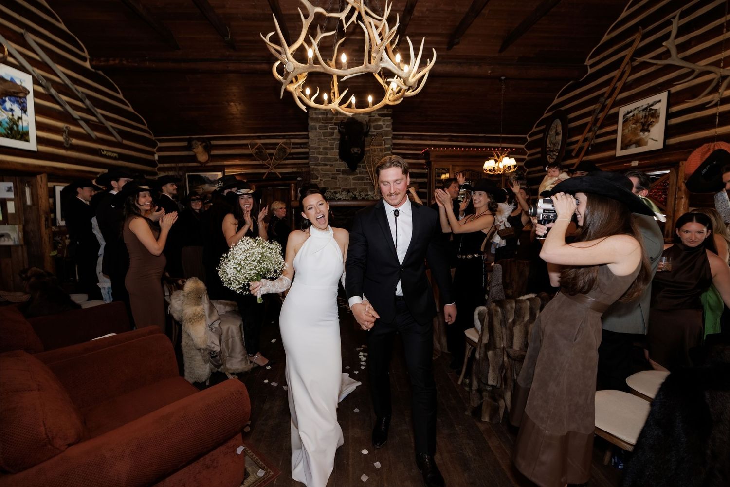 Bride and groom walk hand-in-hand through rustic log cabin wedding venue with antler chandelier and cheering guests.