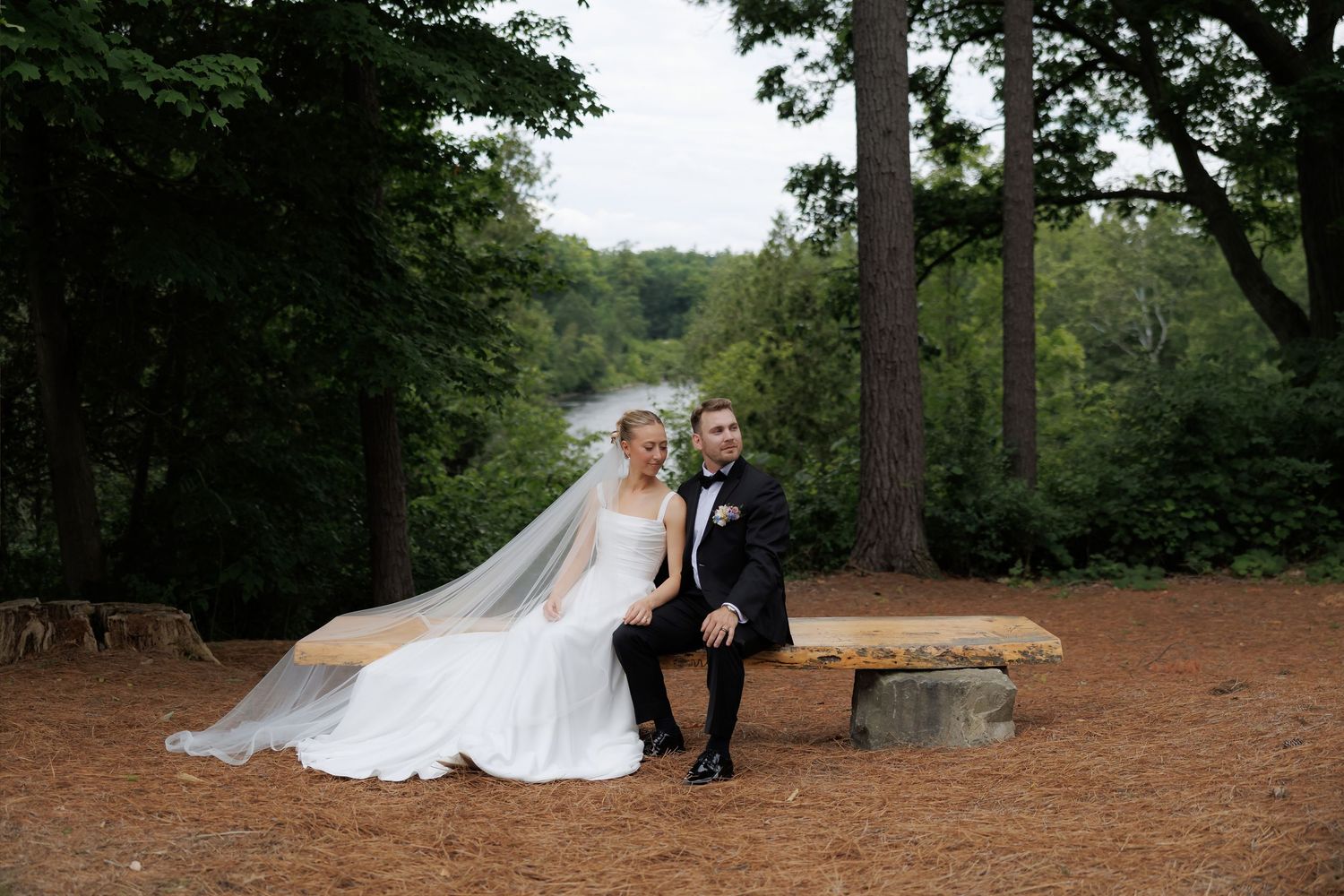 Bride and groom sitting together on a rustic wooden bench surrounded by lush forest and a river.