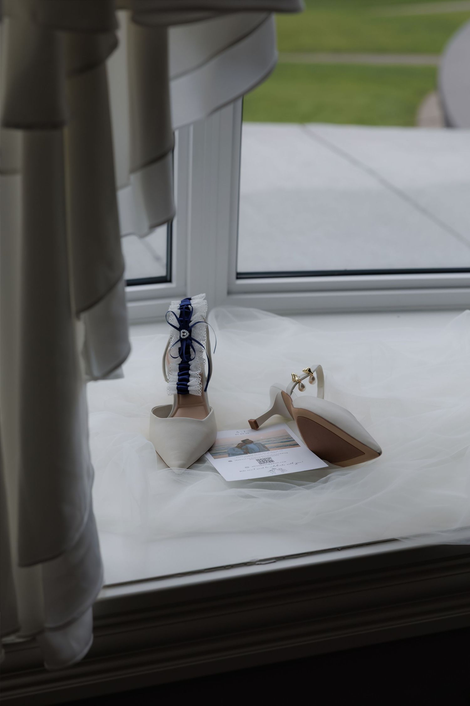 Bridal accessories on a windowsill featuring nude heels with a blue garter and wedding stationery on white fabric.
