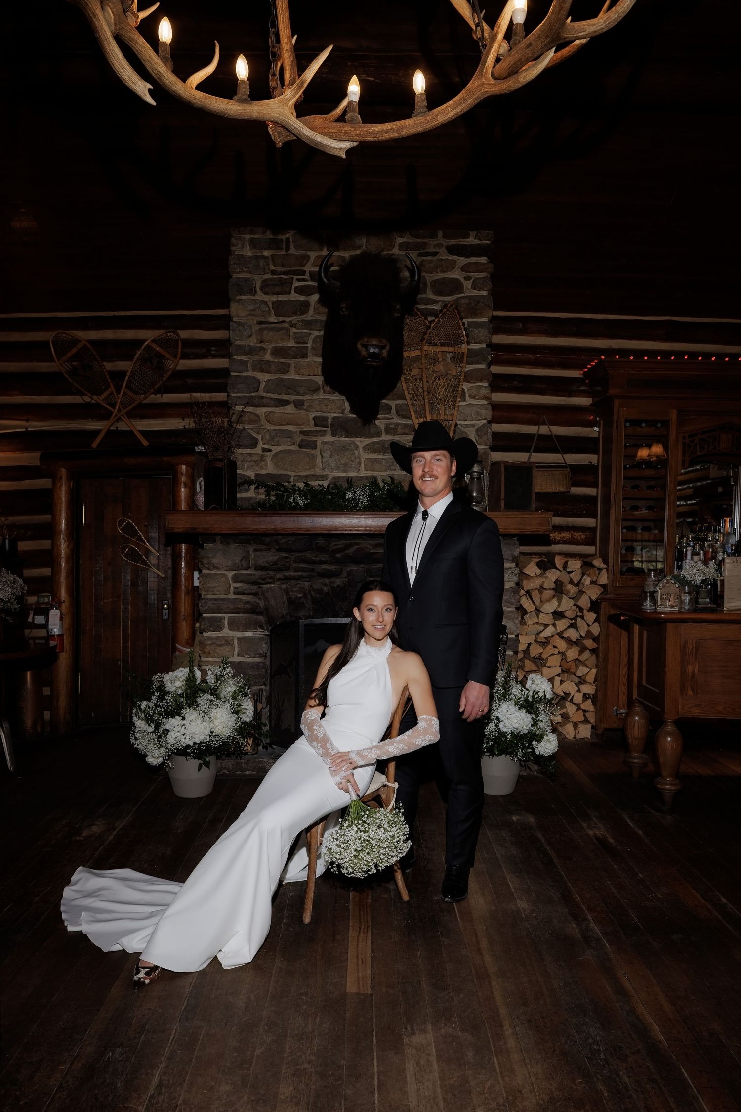 Bride in sleek white gown with lace gloves sits beside groom in black suit and cowboy hat in rustic log cabin.