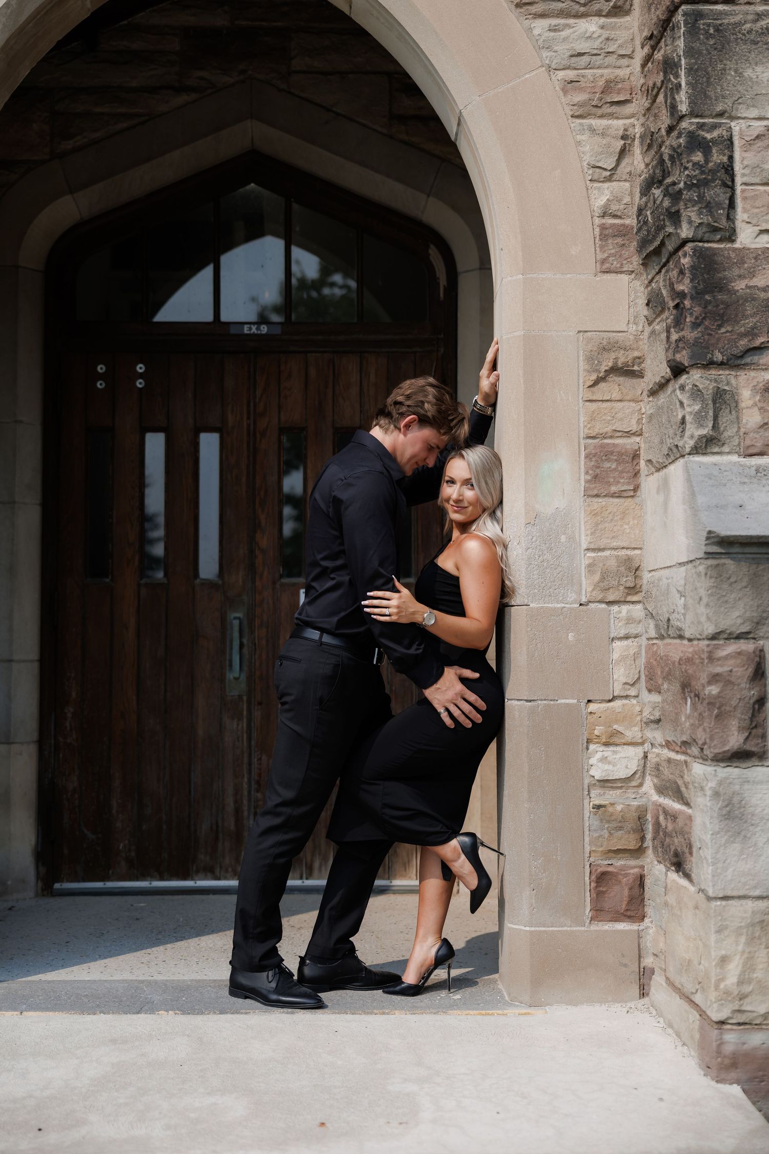 Couple in formal black attire posing romantically in an arched stone doorway.