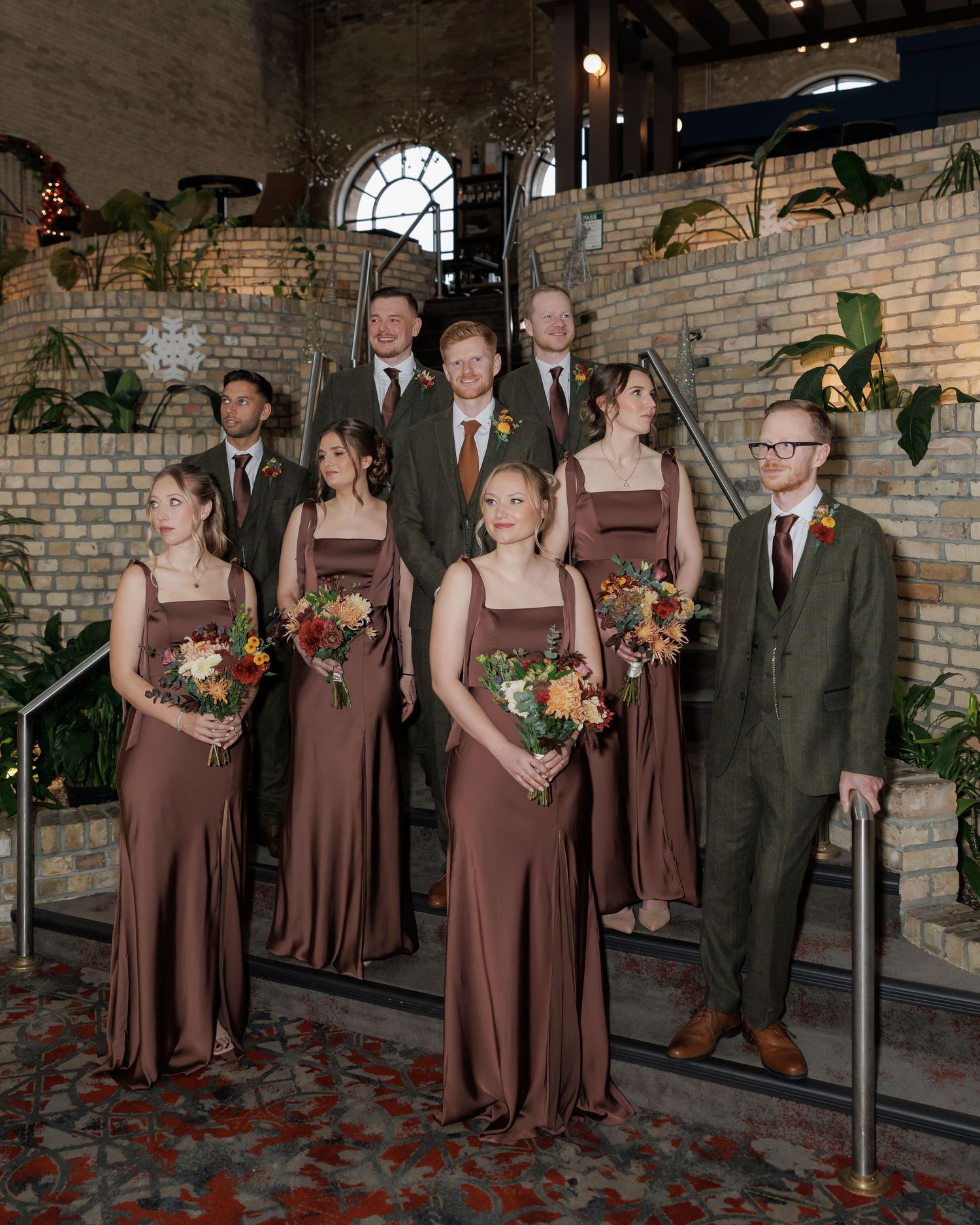 Wedding party poses in brown dresses and suits against exposed brick wall.