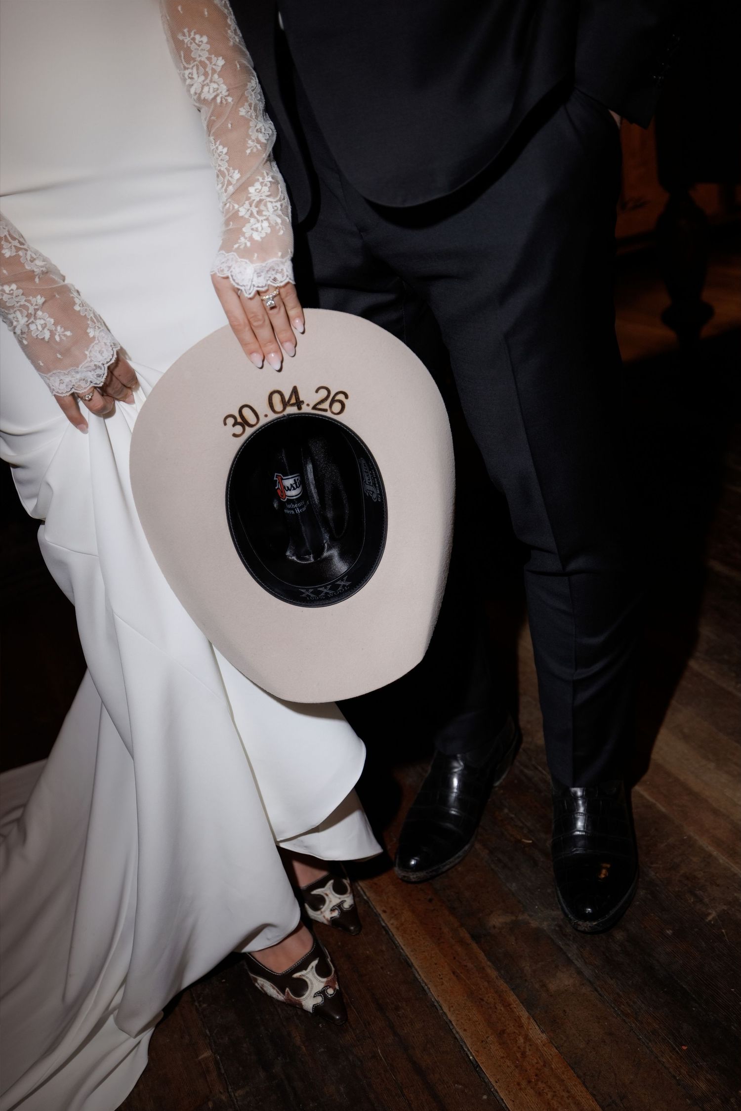 Wedding couple holding a cream cowboy hat engraved with date 30.04.26, bride in lace gown with star-print flats.