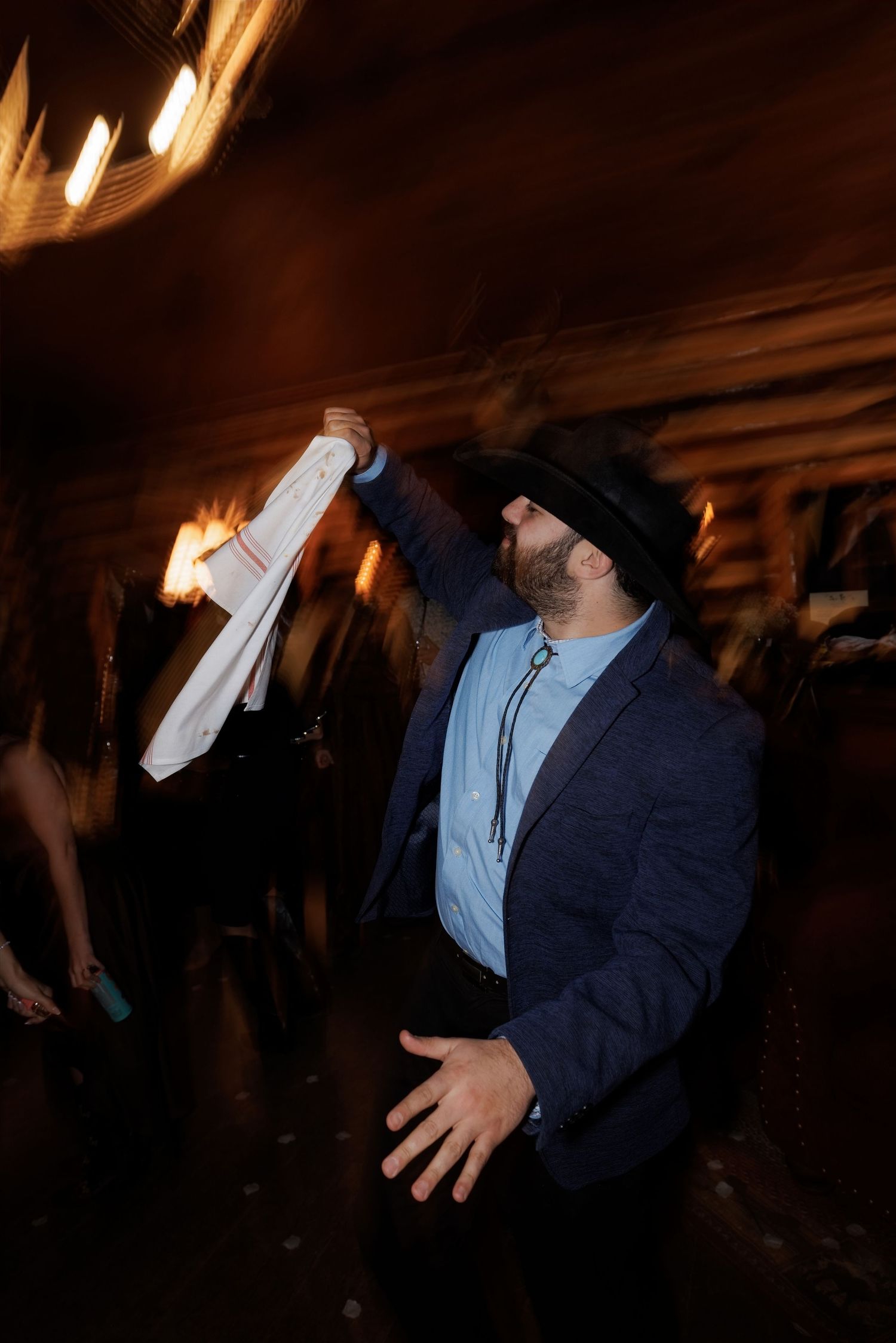 Man in blue blazer and cowboy hat dancing energetically at a dimly lit wedding reception party.