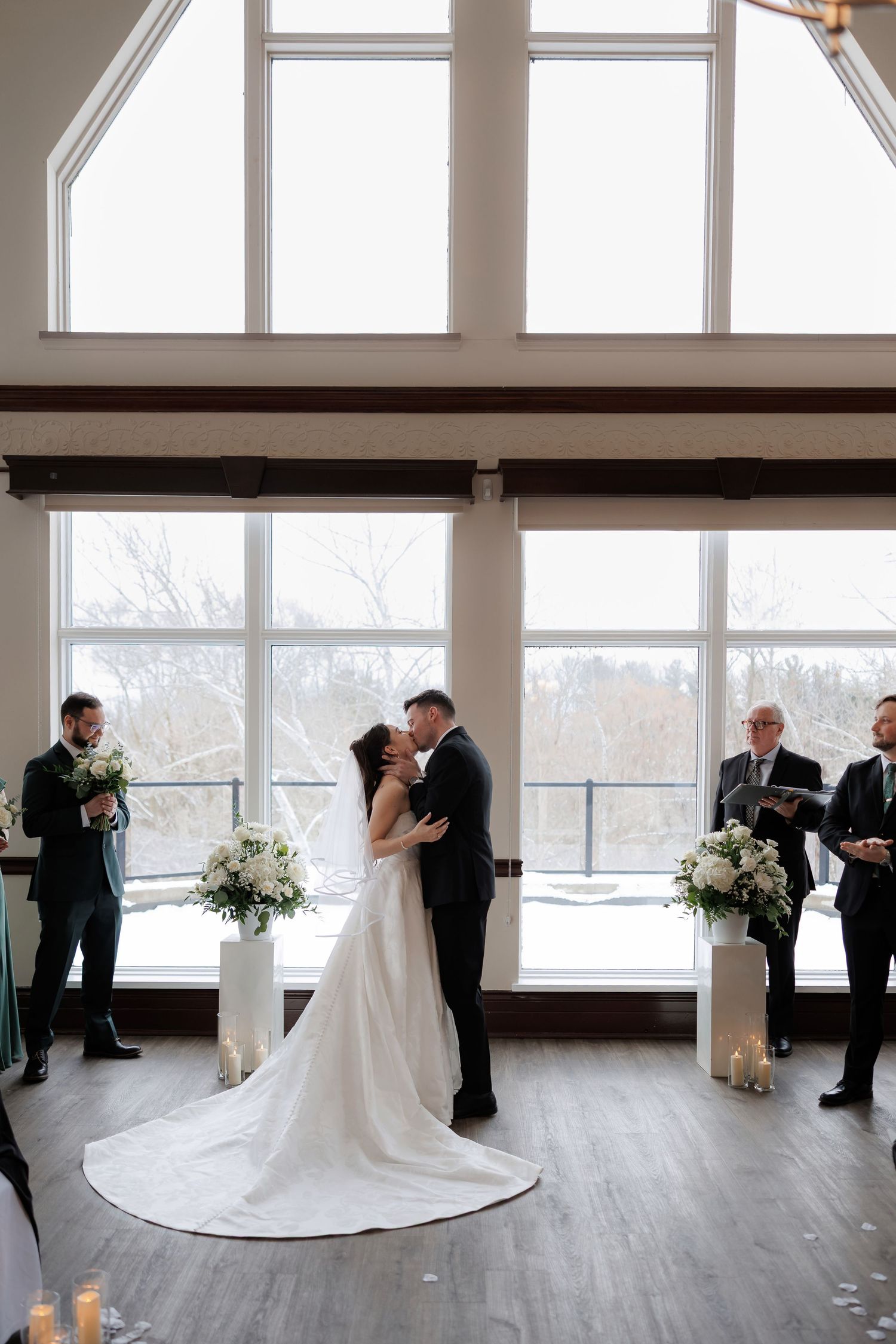 Bride and groom share first kiss at altar with floral arrangements in sunlit venue.