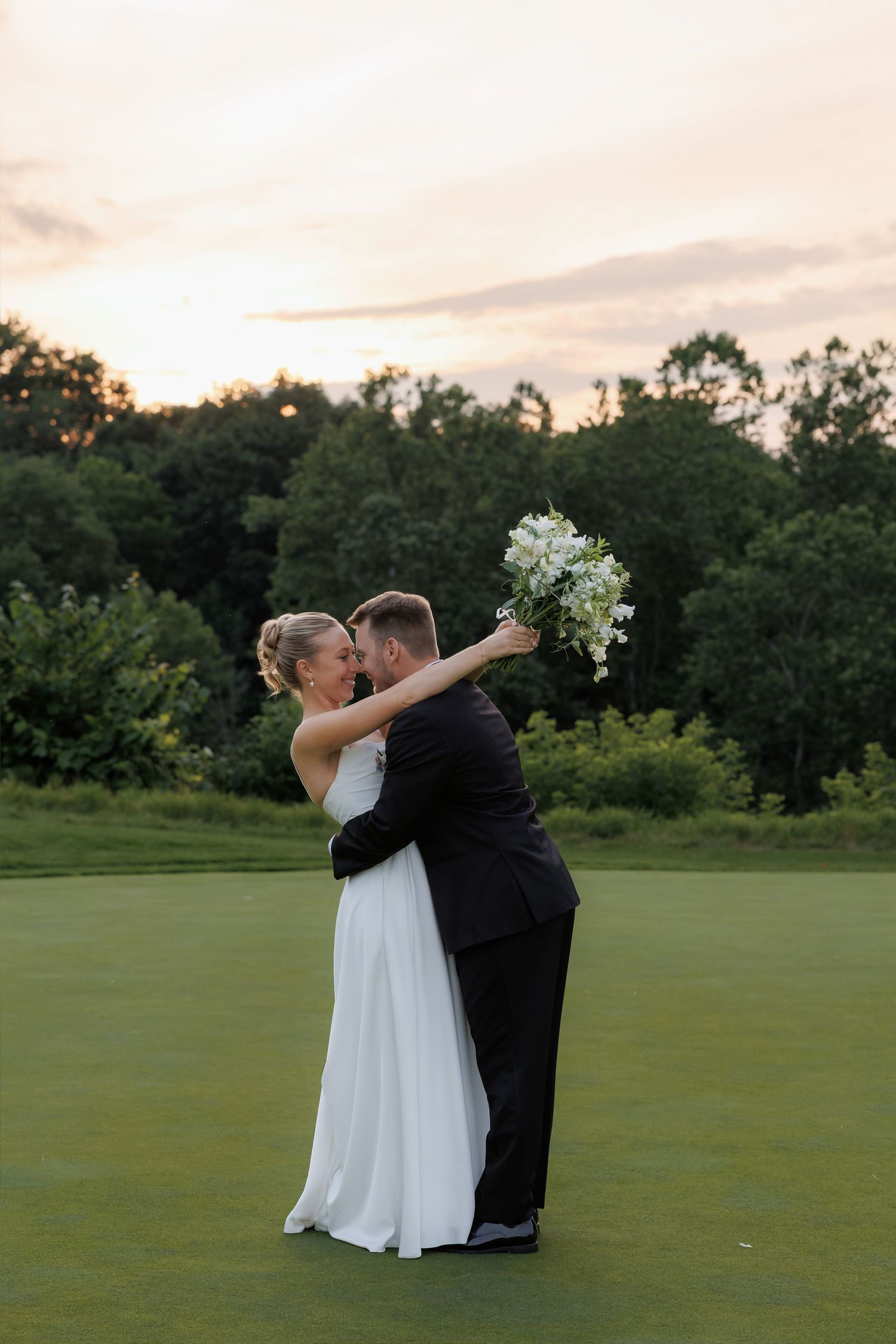 Bride and groom embrace on golf course at sunset, bride holding white bouquet aloft.