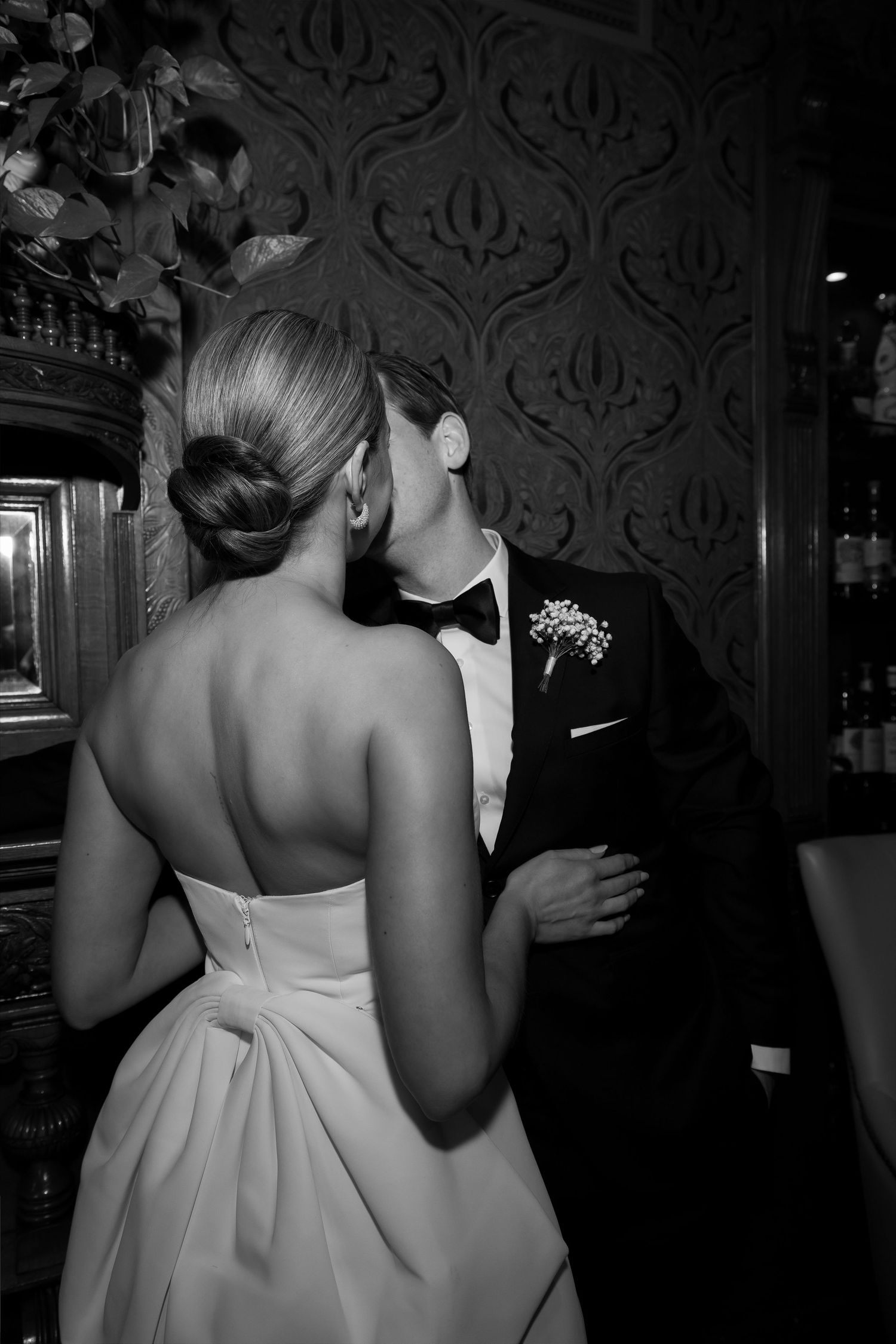 Bride and groom share a passionate kiss against ornate dark wallpaper in elegant black and white portrait.