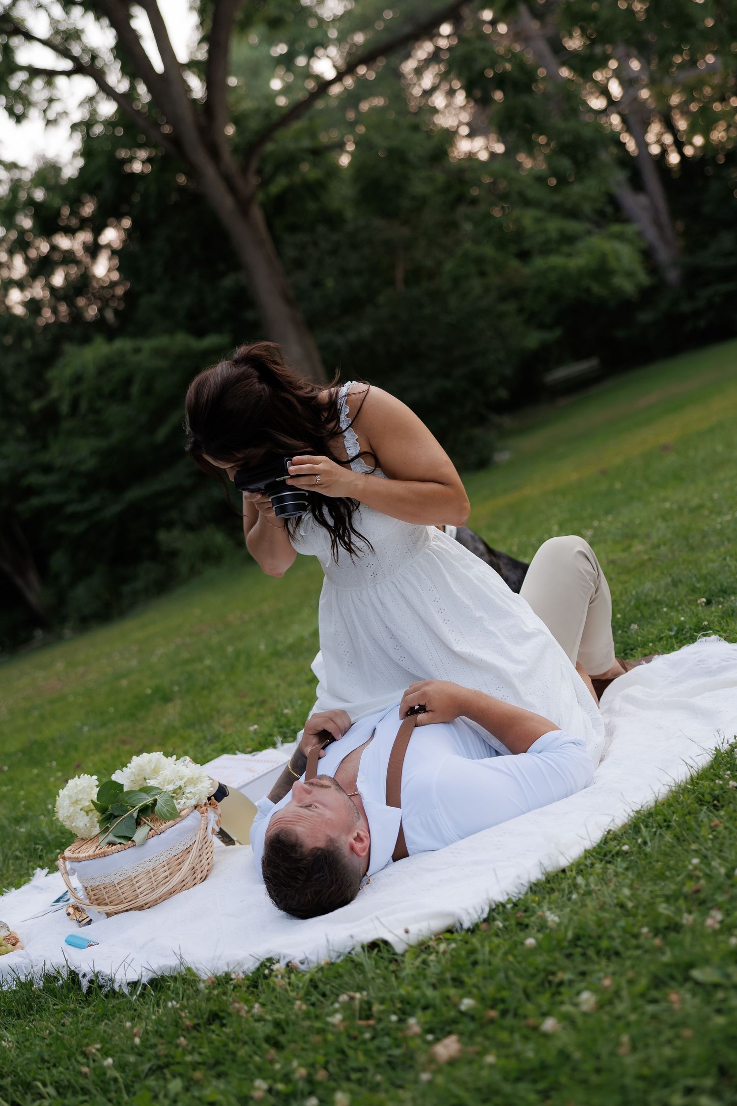 A couple relaxes on a white blanket during a romantic summer picnic in a grassy park at dusk.